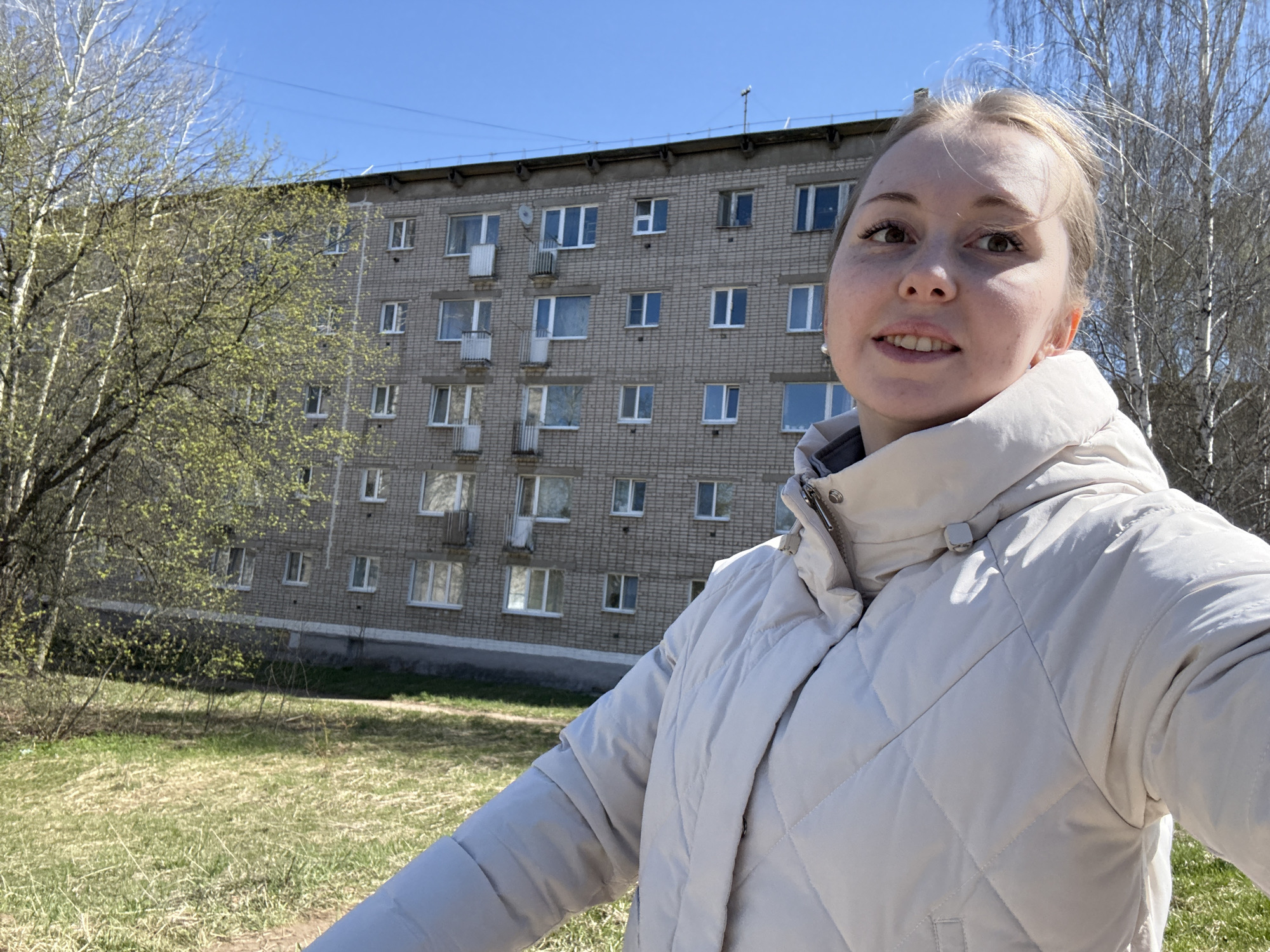 A person is taking a selfie outdoors. The person has fair skin and blonde hair. They are wearing a light-colored puffer jacket. Behind the person is a multi-story brick building with multiple windows and some small balconies. To the left of the building is a tree with budding green leaves, and to the right are bare trees. The ground is covered with patchy green grass and some bare spots. The sky is clear and blue. An antenna is visible on the roof of the building.
