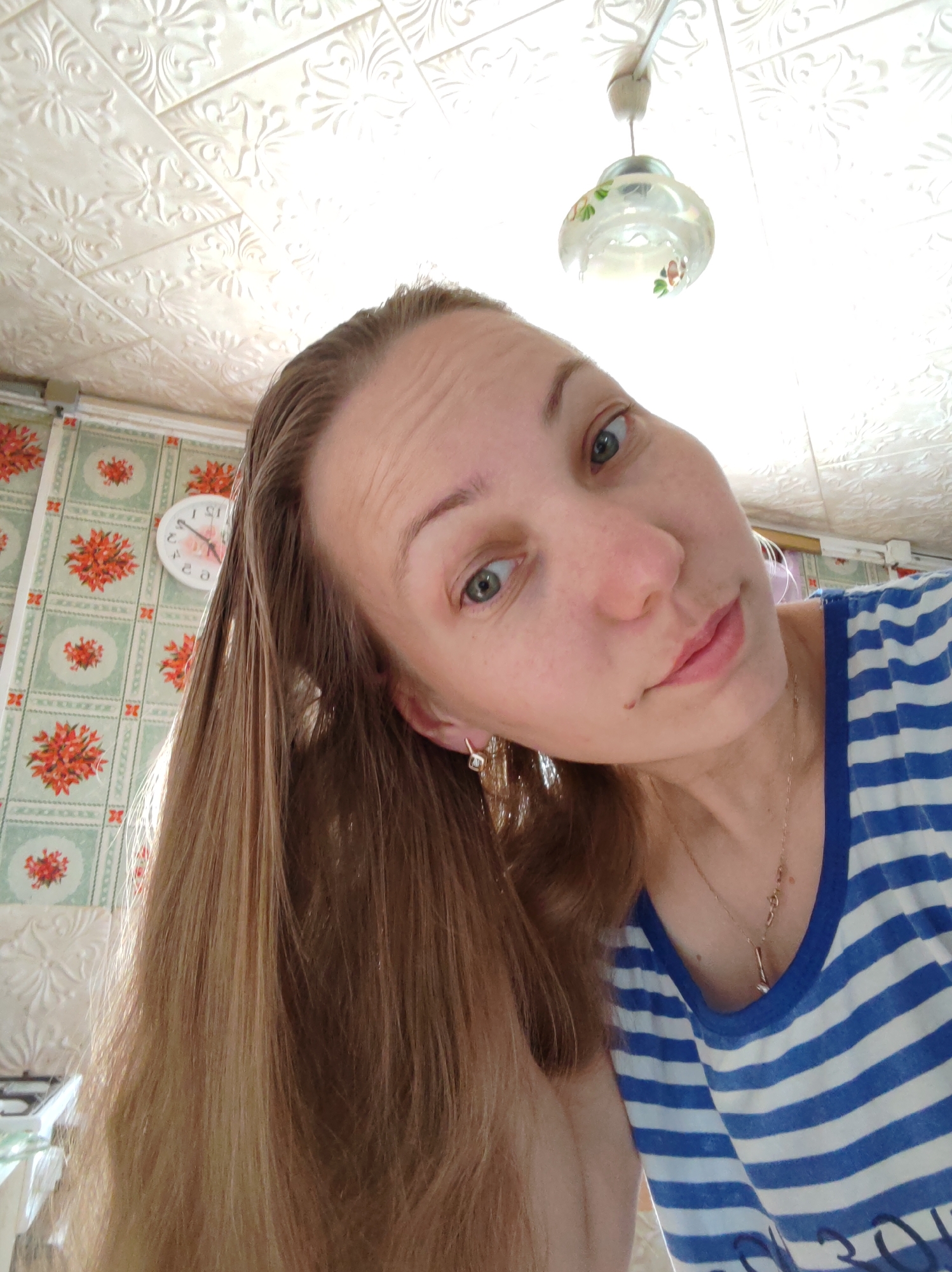 The image is a selfie-style photograph taken from a low angle looking up at a woman. The woman has light skin, light brown hair, and green eyes. She is wearing a blue and white striped shirt, earrings, and a necklace. The background includes a patterned ceiling, wallpaper with a floral design, a clock on the wall, and a light fixture. Based on the geo information it would be located in Чухлэм, Russia.