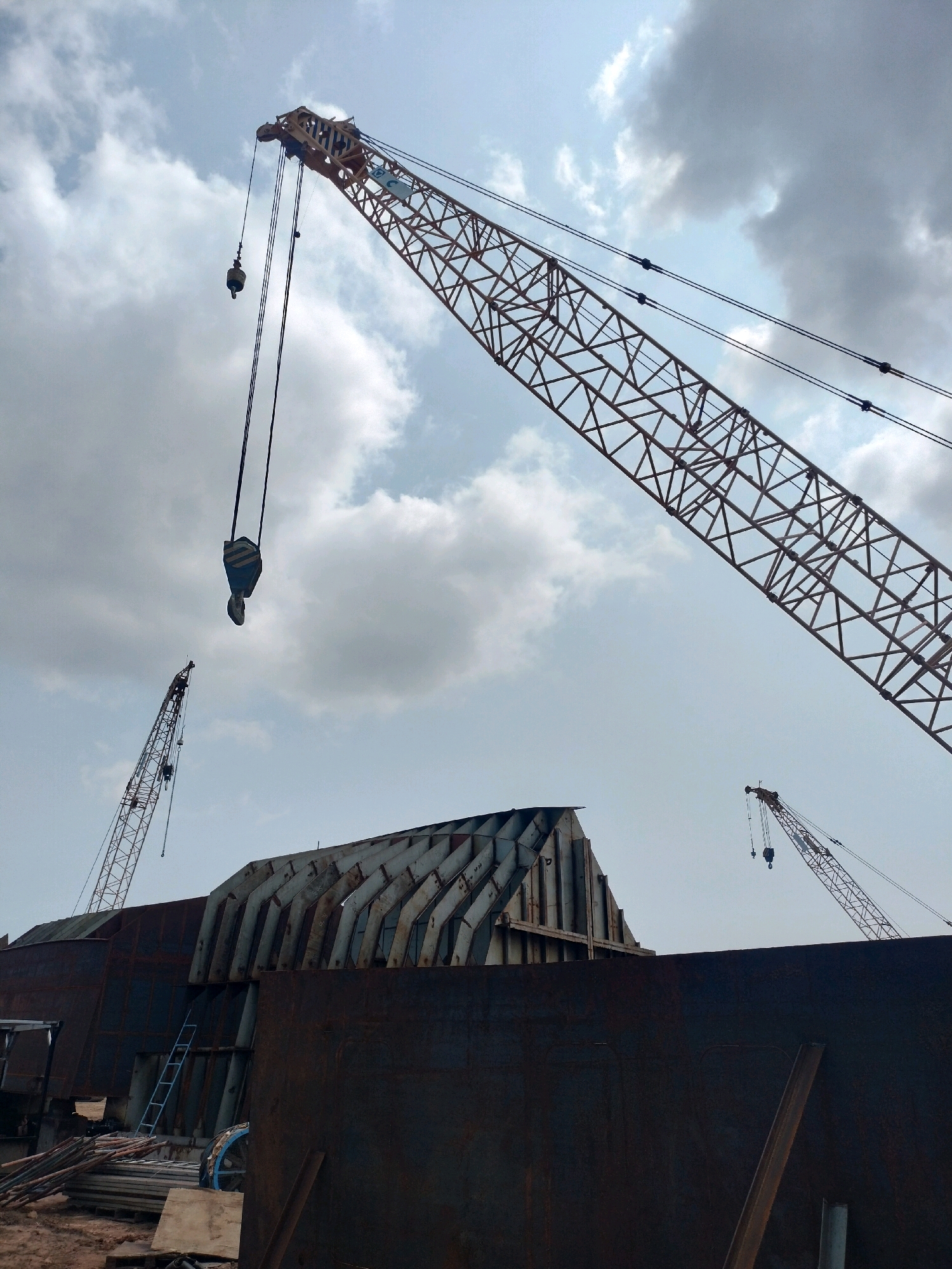 The image depicts a construction site, likely in Batam, Indonesia, characterized by industrial structures and heavy machinery. Dominating the foreground is a massive, dark, curved metal structure, possibly part of a ship or large industrial equipment. To the left, a light blue ladder is affixed to the structure, suggesting ongoing work. Piles of metal pipes and planks are visible on the ground at the base of the structure. In the background, several large cranes are positioned. The most prominent crane, an orange lattice boom crane, extends diagonally across the upper right portion of the frame. A hook and cables hang from its boom. Two smaller cranes are visible further in the distance, also contributing to the industrial landscape under a cloudy sky. The overall impression is one of heavy industrial construction or shipbuilding.