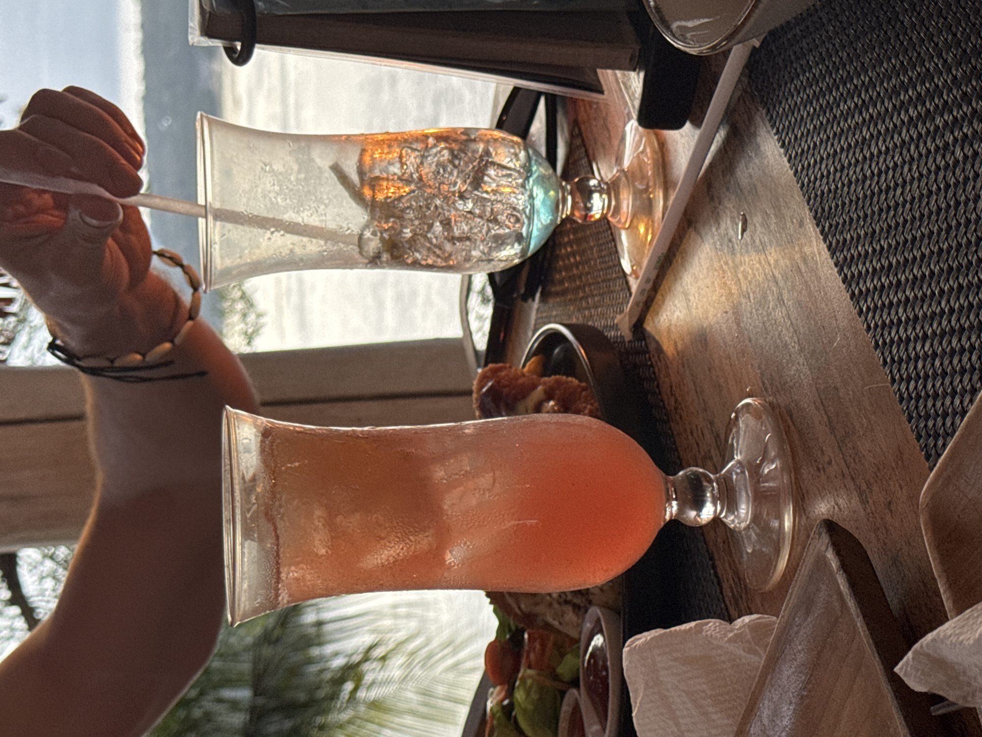 In this media file, the focus is on two tall hurricane glasses filled with beverages, one clear and the other a vibrant pink. A person's arm, adorned with a bracelet made of shells and dark cords, is visible on the left side of the frame, holding a clear straw in the pink drink. The drinks are set on a wooden surface, possibly a table, with a woven placemat partially visible. In the background, out of focus, is a glimpse of what appears to be a body of water and the sky, suggesting an outdoor setting, likely a restaurant or bar by the coast. The lighting indicates it is likely daytime, possibly late afternoon given the warm glow on the drinks and table.
The scene captures a moment of relaxation and enjoyment, with the drinks suggesting a leisurely activity. The presence of the drinks and the casual attire implied by the bracelet hint at a vacation or a relaxed outing. The location context of Nusa Penida, Indonesia, further reinforces this idea, evoking images of tropical beaches and holiday destinations. The overall mood is one of tranquility and indulgence. There is no visible text in the media file.