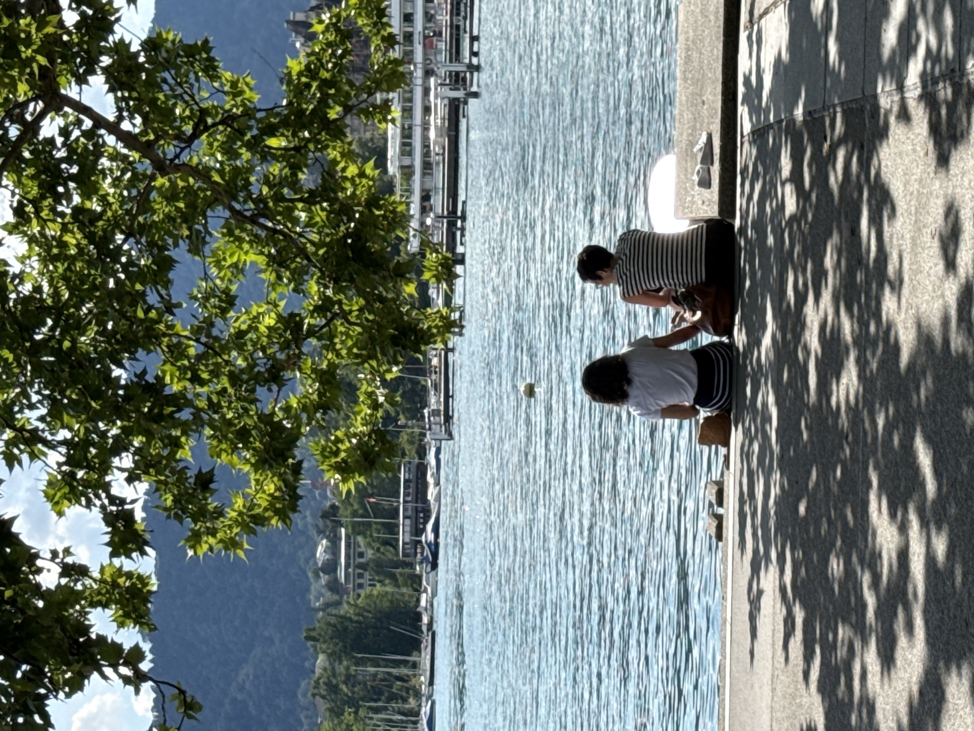 Two individuals are seated on a concrete embankment by a body of water, viewed from behind. The person on the left has dark hair and is wearing a white t-shirt with a black and white striped skirt. The person on the right has short dark hair and is wearing a black and white striped t-shirt and dark pants. A brown bag is placed between them. Two light-colored shoes are on a stone surface to the right of the individuals. The water is blue and appears slightly rippled. In the background, there are several boats docked or on the water, and a large white passenger boat is visible further out. Trees with green leaves are in the foreground, casting shadows on the embankment. Beyond the water and boats, a hilly landscape with vegetation and buildings can be seen, under a partly cloudy blue sky. This scene is consistent with the lakeside promenade in Zurich, Switzerland.