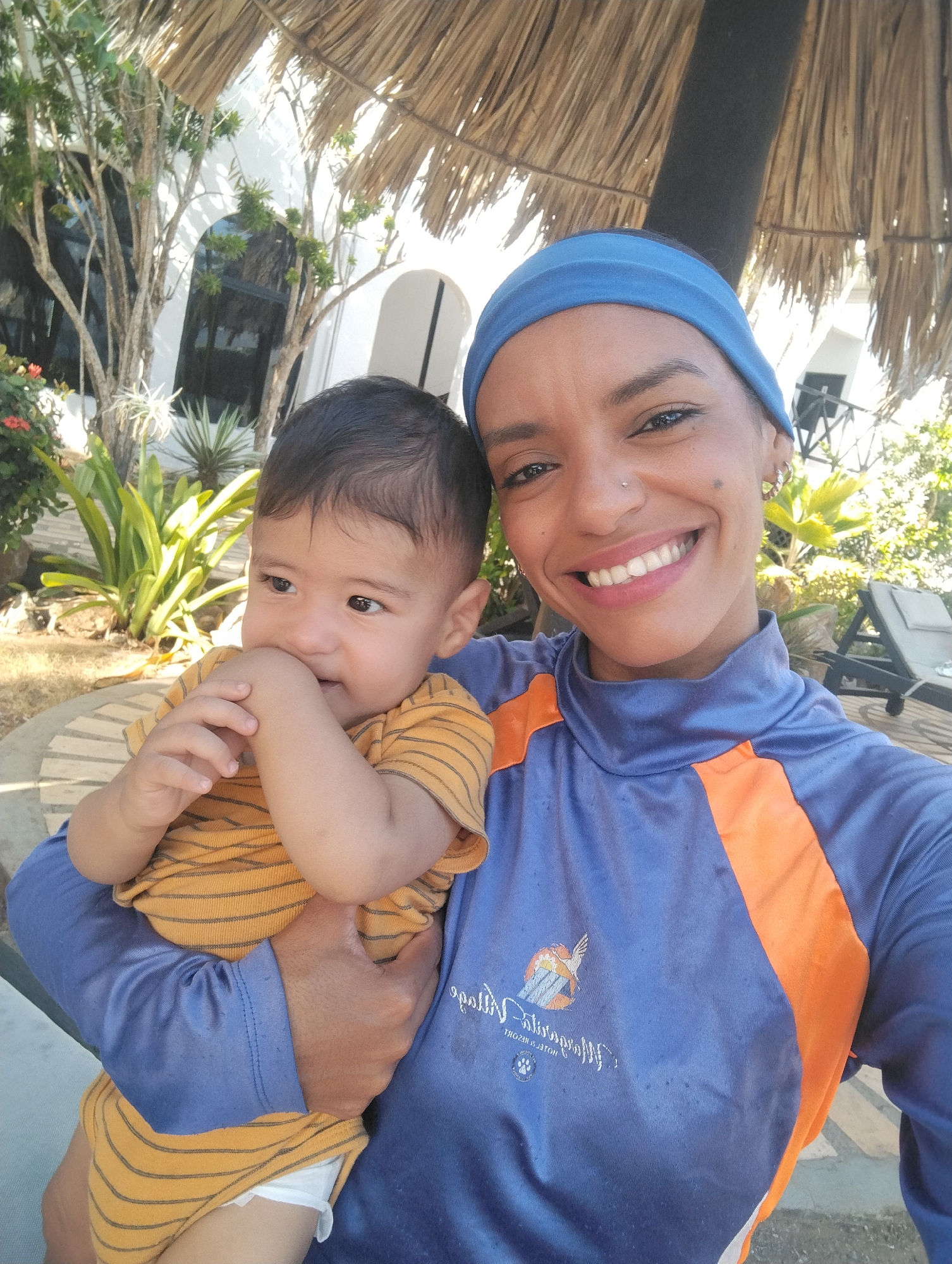 The image shows a woman holding a baby. The woman is wearing a blue headband and a blue and orange long-sleeved shirt with a logo. She is smiling broadly, revealing her teeth. The baby is wearing a yellow and brown striped shirt and appears to be chewing on his hand.
They are standing outdoors, possibly on a patio or near a garden, as indicated by the presence of tropical plants, a white building with arched windows, a thatched roof structure, and lounge chairs.
The logo on the woman's shirt appears to be "Hesperia Isla Margarita" and "HOTEL & RESORT". This suggests that the location is likely the Hesperia Isla Margarita hotel in Porlamar, Venezuela.