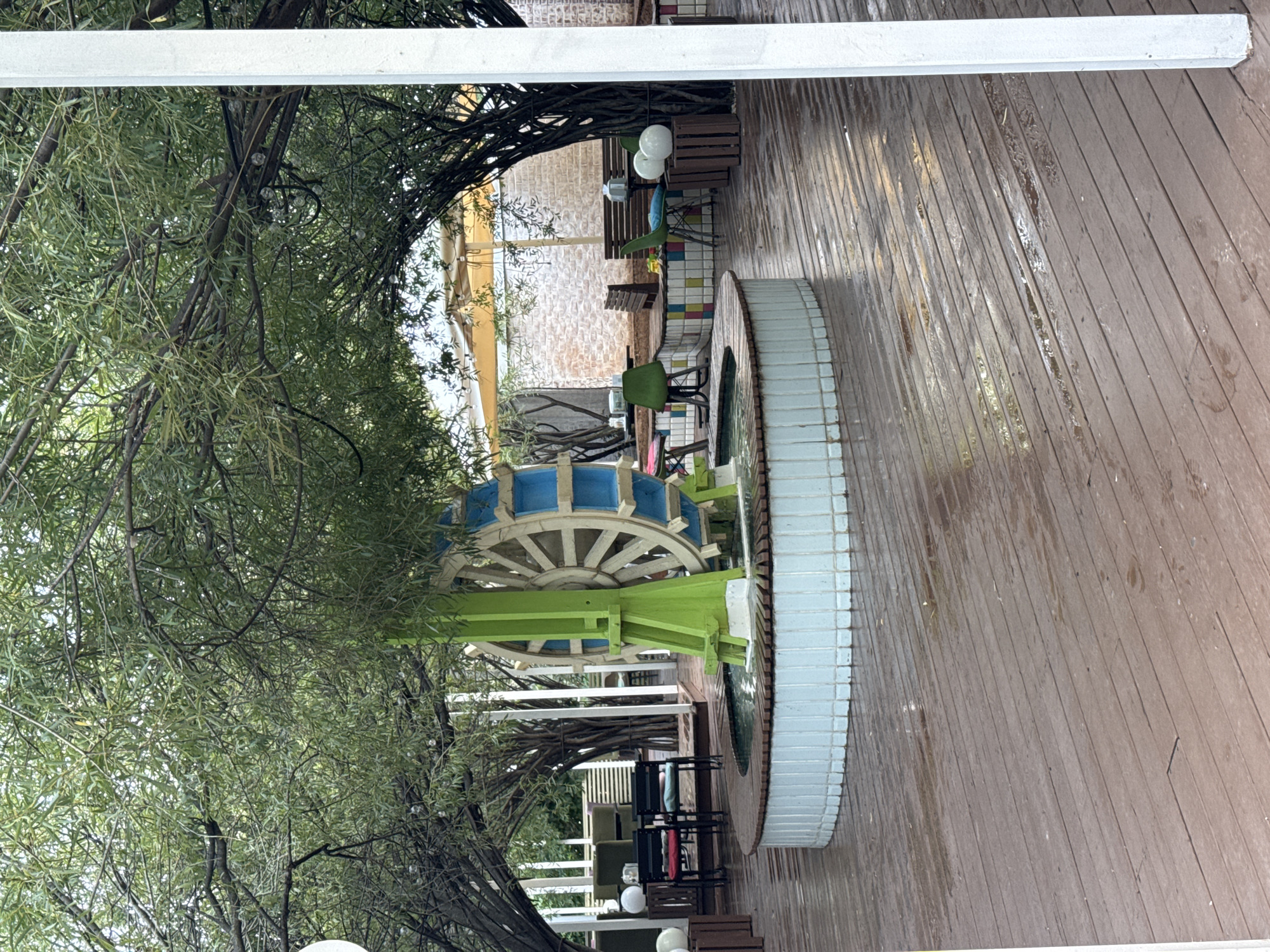 The image depicts an outdoor seating area, likely a cafe or restaurant, with a central water feature. The area is covered by a canopy of trees with dense foliage, creating a natural, shaded ambiance. In the foreground, a wooden deck, wet from recent rain, slopes gently away from the viewer.
Dominating the center of the frame is a circular, tiled water basin with a wooden rim. Within the basin, a decorative water wheel is prominently displayed. The water wheel has a pale wooden exterior with blue accents and is supported by a bright green, angular structure. The water in the basin appears calm.
Surrounding the water feature, further seating arrangements are visible. To the left, two black chairs with red and blue cushions are positioned at a small table. Further back, other seating areas with tables and chairs, some with benches, are scattered throughout the space. Some tables have white spherical decorations on them.
In the background, a brick wall is visible, and above it, a pale yellow awning provides additional shelter. The overall impression is of a serene, outdoor space designed for relaxation, with a playful, decorative element in the water wheel. The image does not contain any visible people or specific landmarks that would place it in Ufa, Russia.
