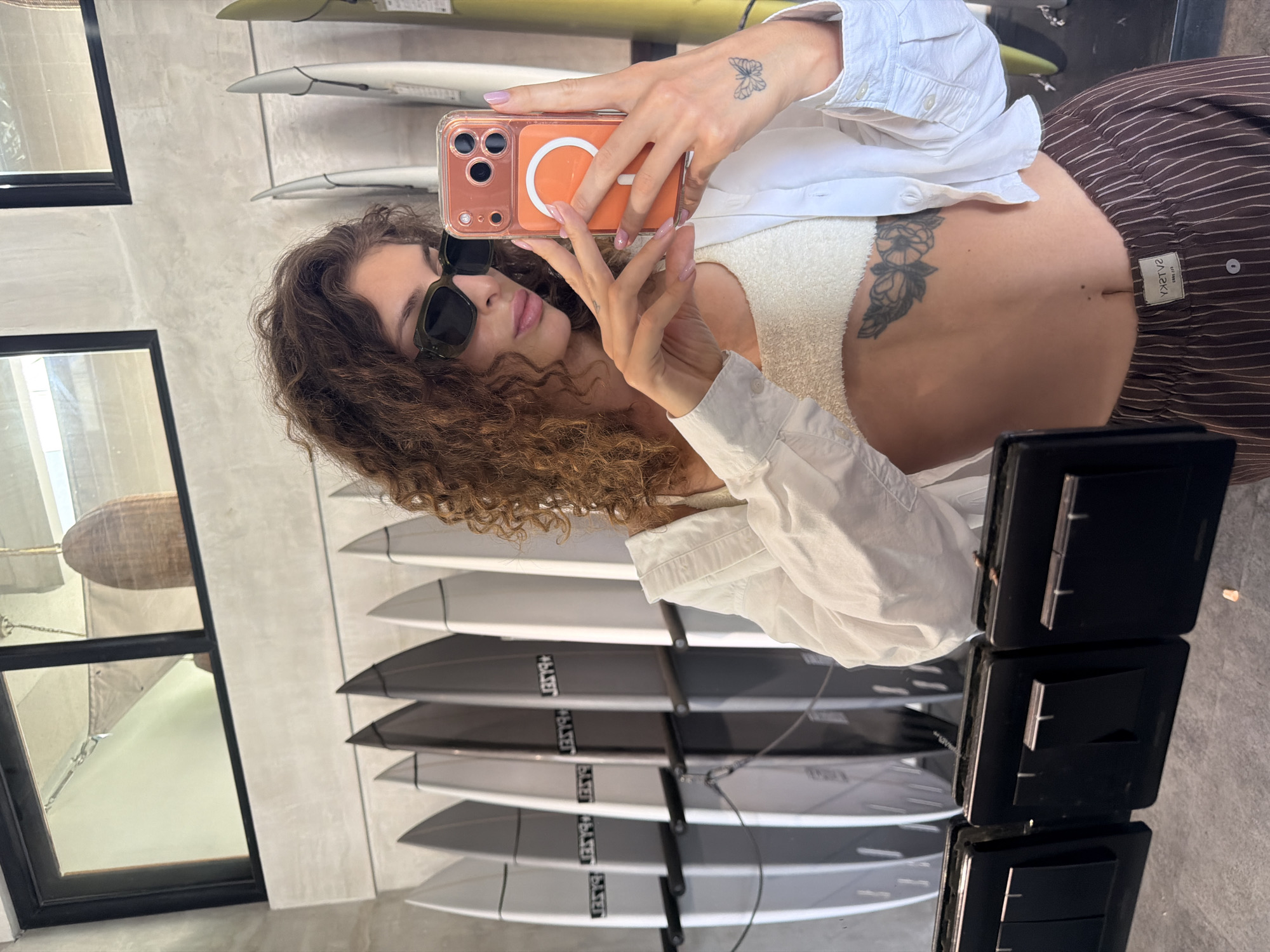A woman is taking a selfie in what appears to be a retail space, possibly a surf shop, in North Kuta, Indonesia. The woman has curly hair and is wearing sunglasses, a white shirt, and striped pants. She's holding an iPhone with an orange case, and the reflection shows several surfboards displayed on a rack behind her. Visible on her body are tattoos of a butterfly on her wrist and a floral design near her torso. The background shows a window with black frames and a glimpse of an interior space with what looks like woven baskets or decorations. Also visible are black light switches on the right side.