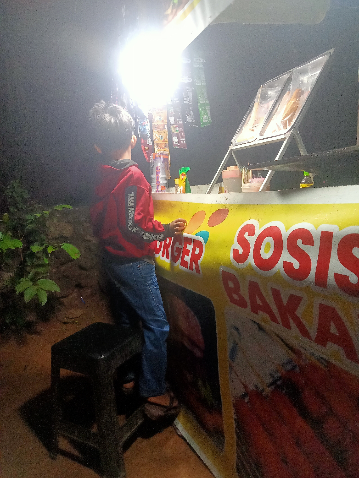 A young boy in a red hoodie and blue jeans stands on a black plastic stool in front of a food stall. The stall has a bright yellow side with red lettering that reads "SOSIS BAKAR," and the word "BURGER" is partially visible. There are various items displayed on shelves above the stall, including packaged snacks and drinks. To the left of the stall, there is lush green foliage. The scene is illuminated by a bright light source above the stall, creating a dramatic contrast with the dark surroundings. The location is identified as Tanggaran, Indonesia.