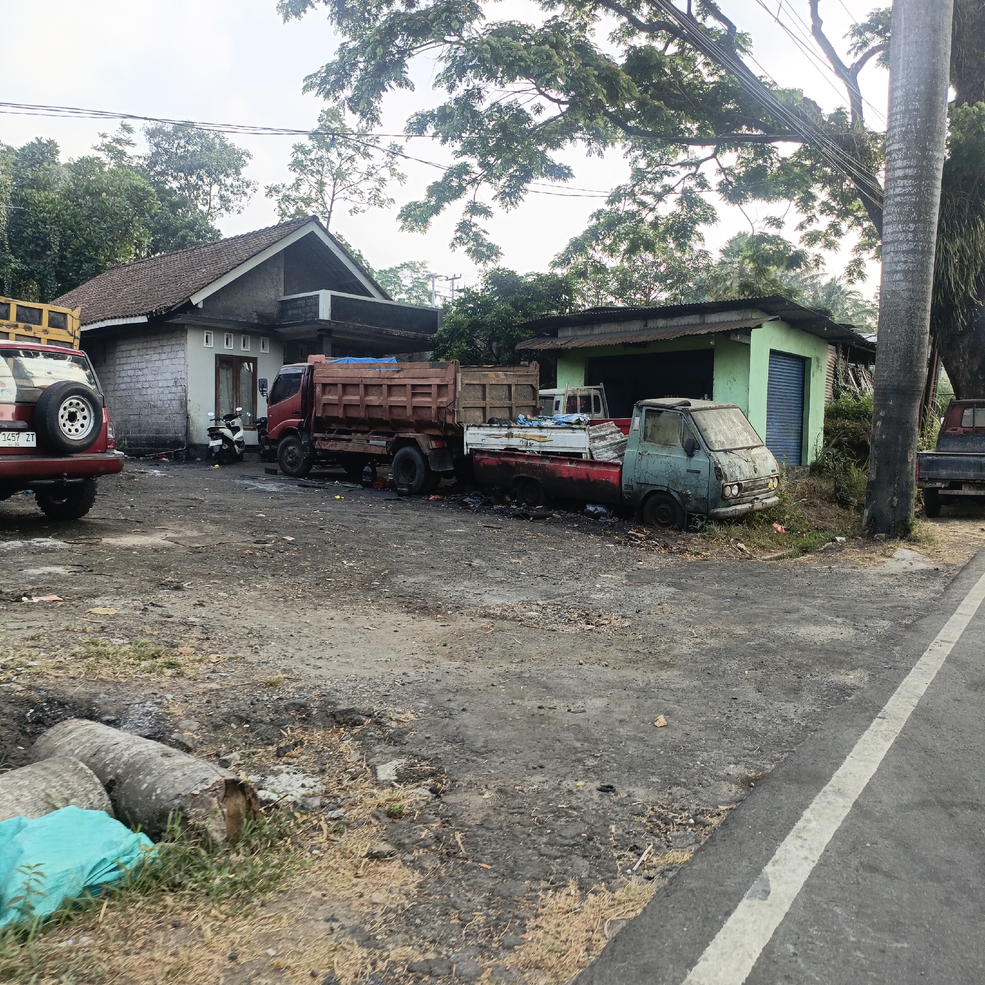 The image depicts a dusty, unpaved lot adjacent to a road in Salatiga, Indonesia. Several vehicles are parked on the lot, some appearing weathered and potentially abandoned. In the foreground, a red SUV with a spare tire mounted on the back is visible. Behind it, a red dump truck is parked. To the right of the dump truck is a small, teal-colored truck, also showing signs of age. A low-lying green building with a corrugated metal roof is situated behind the teal truck. A residential building with a tiled roof and light-colored walls is visible further back. Trees with dense foliage are scattered throughout the scene, and power lines are strung overhead. The ground is covered in dirt, gravel, and debris. A portion of an asphalt road with a white line marking is visible on the right side of the image. The lighting suggests it is daytime, possibly early morning or late afternoon.