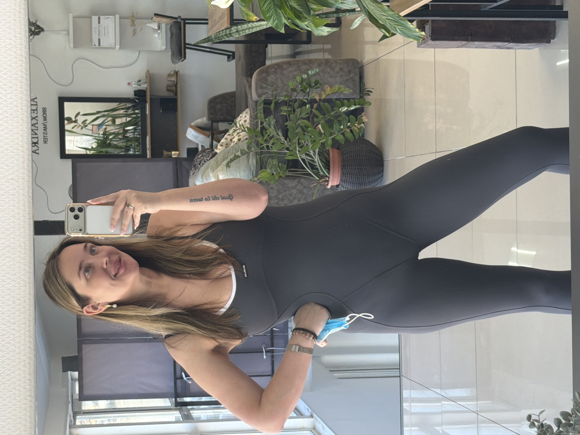 A woman is captured in a mirror selfie, extending her left arm to hold a silver-colored smartphone with three rear cameras. She has long, light brown hair, is smiling with her tongue slightly out, and wears a gold earring with a white pearl and a delicate necklace. Her attire is a form-fitting, dark gray or black one-piece athletic or casual jumpsuit with a white trim visible at the neckline. Her left wrist displays a silver-toned watch and several beaded bracelets. In her right hand, she holds a light blue disposable face mask.
A tattoo is visible on her right forearm, reading "Quod aliis licet tibi non licet." The background, visible in the mirror's reflection, suggests an indoor commercial space, possibly a beauty salon or studio. A white sign on the wall behind her is reflected as "ALEXANDRA BROW MASTER." Other elements in the reflection include a framed mirror, various potted plants, shelves with items, and upholstered furniture. The flooring appears to be light-colored tiles. The lighting is bright, indicating daytime. The woman appears cheerful and playful.