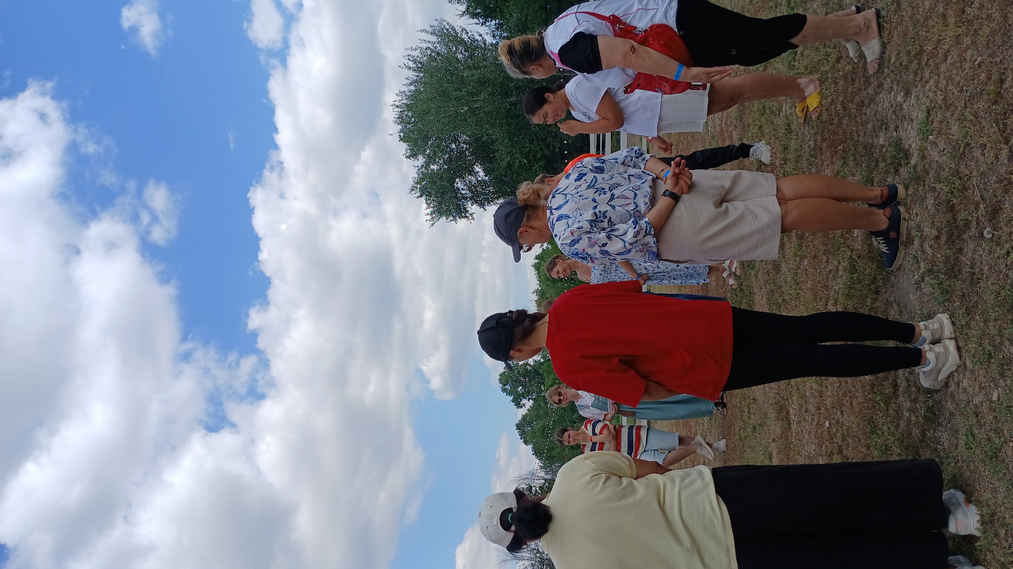 A group of people are standing in a circle in a dry, grassy field under a partly cloudy sky. The background consists of trees and a white fence. The people are dressed in casual clothing. One person in the foreground, facing away from the camera, is wearing a yellow shirt and black pants. To their right, a person in a red shirt and black leggings is also facing away. Another person to the right is wearing a patterned blue and white shirt and beige shorts. To the far right, a person in a white top and black pants with a red bag slung over their shoulder is visible. In the middle of the circle, several other individuals are standing, including one woman in a blue dress and sunglasses, and another in a striped shirt and shorts. The overall impression is that of a casual outdoor gathering in a rural or park-like setting, possibly in the city of Derköl, Kazakhstan.