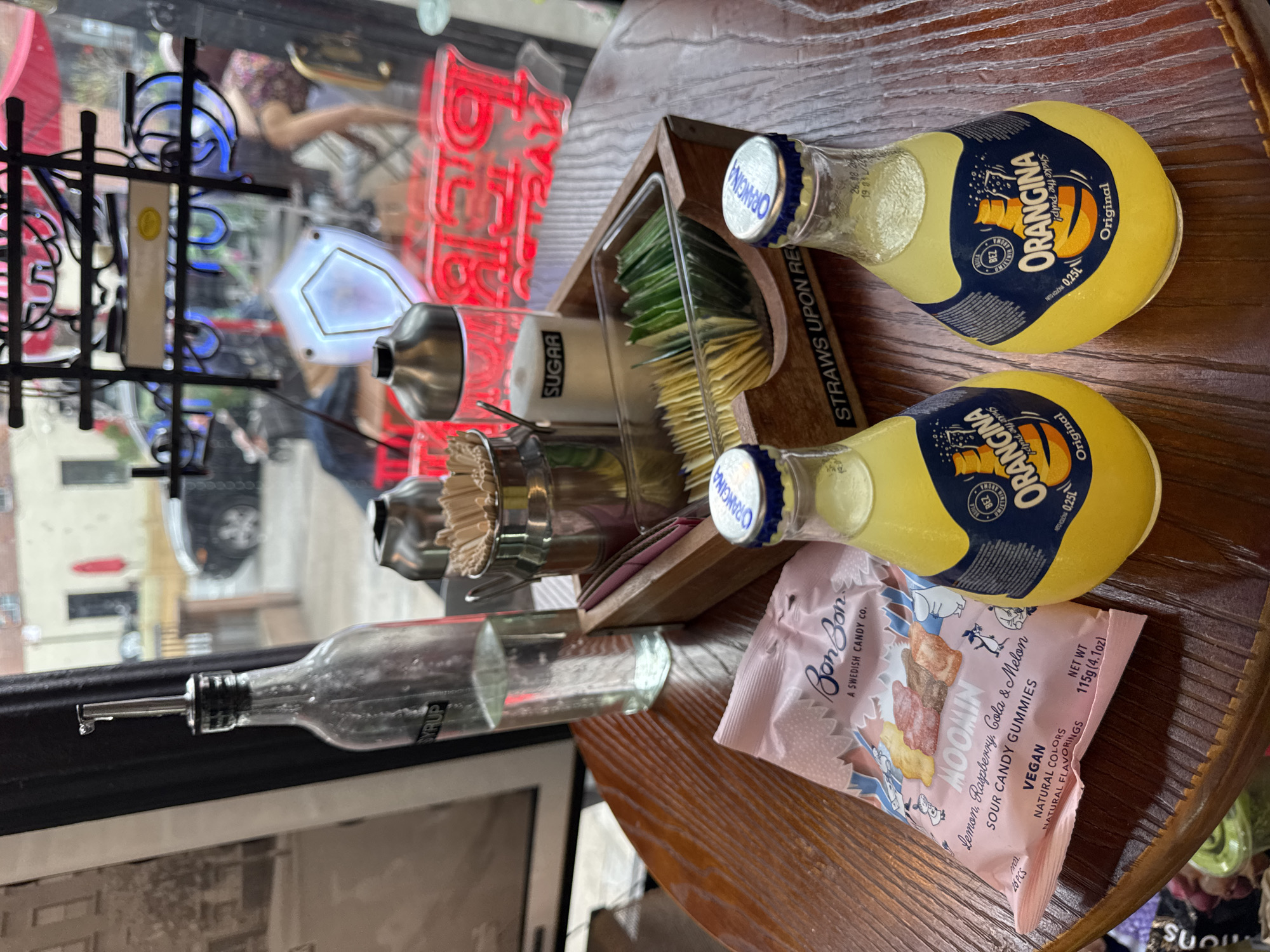The image depicts a tabletop scene, likely inside a café or restaurant. Two small glass bottles of Orangina are positioned on the right. Near them is a pink packet of Bon Bon brand Moomin-themed sour candy gummies. Adjacent to the Orangina is a wooden tray containing sugar packets and straws. To the left of the tray is a glass container holding toothpicks and a bottle of syrup with a spout. The background shows reflections in a window, including neon signs and street scenes. The reflections suggest the location is likely in New York City, United States of America. The table itself appears to be made of wood.