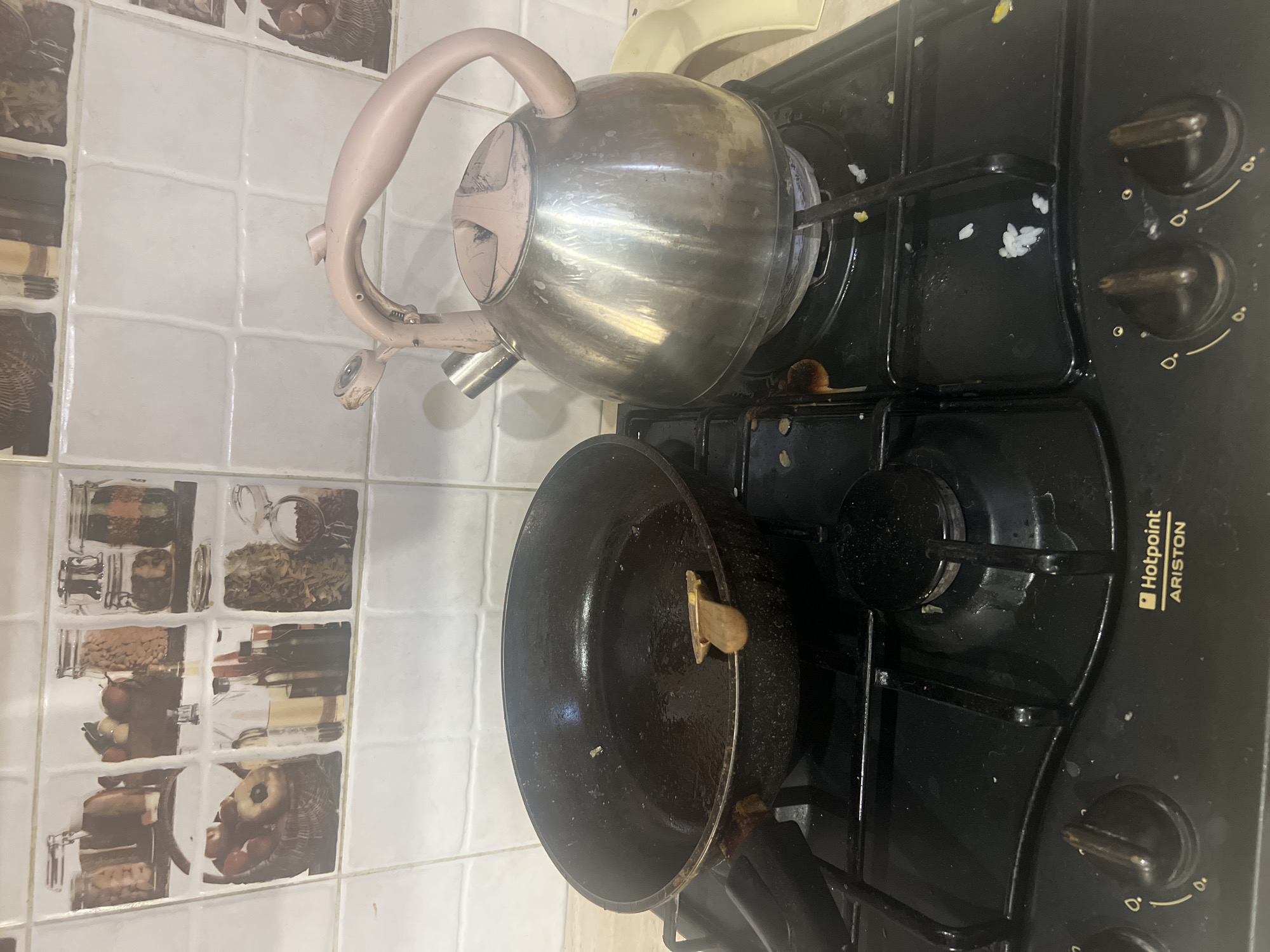 The media file shows a close-up, vertically oriented shot of a stovetop in a kitchen. The subjects are a metal kettle with a light pink handle, and a black frying pan. The kettle is positioned on the left side of the stovetop, while the frying pan is in the center. The stovetop itself is black with metal grates and two visible control knobs. There is some scattered food debris, appearing to be grains of rice, on the stovetop. The background consists of tiled walls with decorative images of jars containing food items like spices and grains, and some baskets of fruit.
The scene appears to be in a domestic setting, likely a kitchen, given the presence of the stovetop, kettle, and frying pan. The lighting suggests it could be daytime, but there are no strong indicators of the time of day or weather. The overall impression is of a functional kitchen space.
The text visible on the stovetop is "Hotpoint ARISTON". There are no people visible in the media file, and no specific activity or event is clearly depicted, beyond the potential preparation of food. The focus is on the kitchen appliances and the immediate surroundings.
