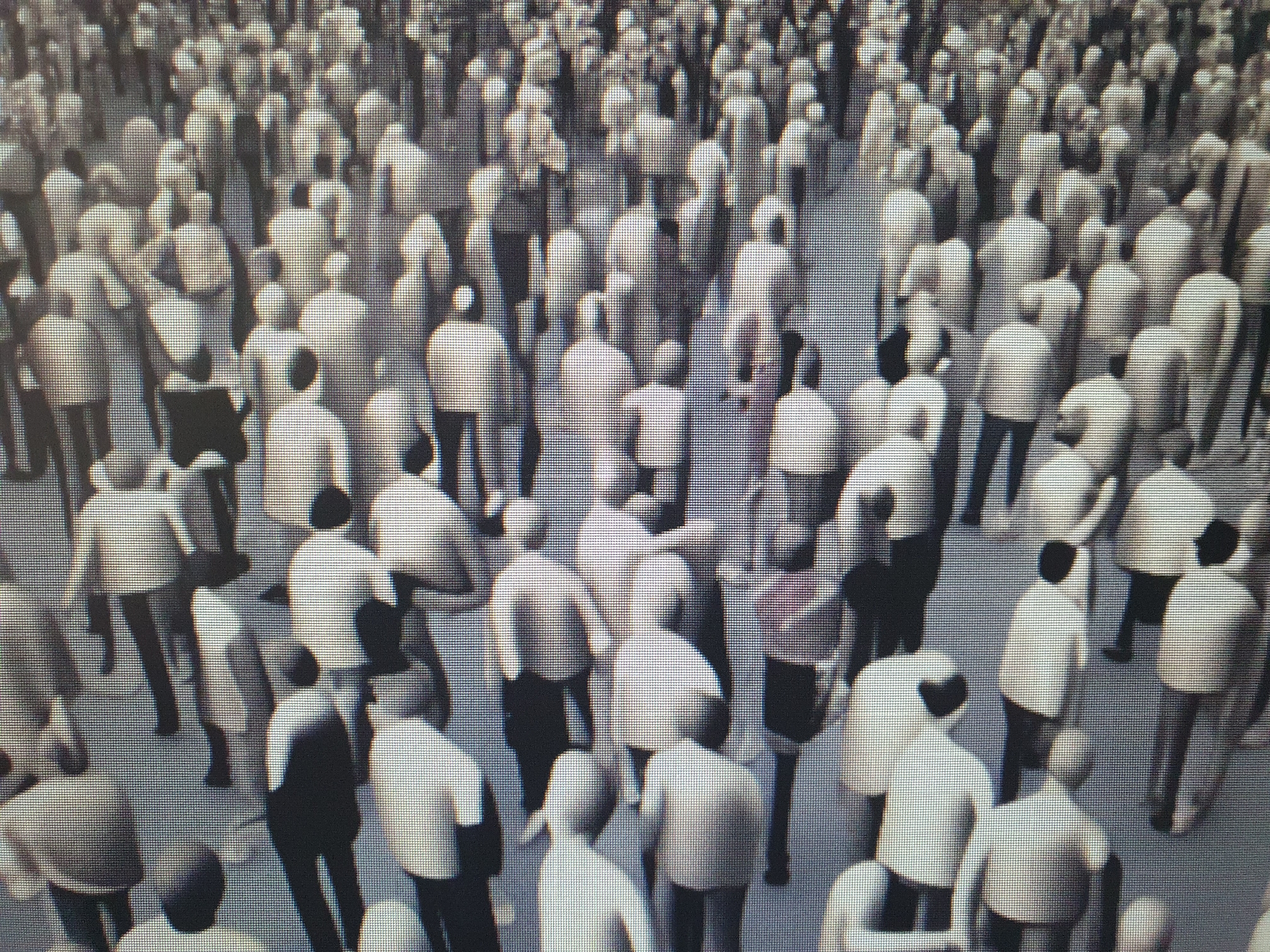The image depicts a high-angle, somewhat blurry view of a dense crowd of people. The individuals are mostly standing or walking in various directions, creating a sense of a public gathering or busy thoroughfare. They are dressed in light-colored tops and dark bottoms, a common clothing style. The scene appears to be outdoors, with a textured, grid-like ground surface. The overall impression is of a large group of individuals in a public space, possibly a city square or market. There is no identifiable signage or architectural detail to pinpoint a specific location within Tashkent, Uzbekistan.