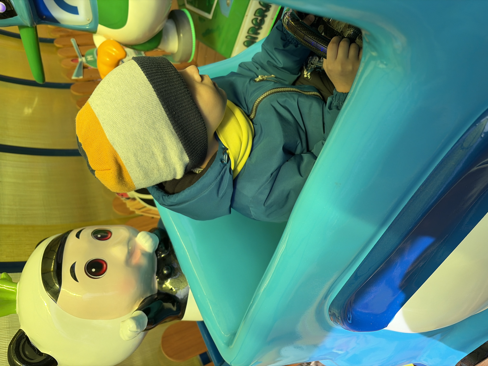 The image displays a young child seated in a vibrant turquoise-blue kiddie ride, likely in an indoor amusement area or play zone. The scene is brightly lit, suggesting artificial indoor lighting.
The child, a primary subject, is seen from the chest up, wearing a multi-striped beanie with white, yellow, and dark grey sections, and a blue zip-up jacket over a yellow garment. Their hands are placed on a dark steering wheel, indicating engagement with the ride. The child's face is visible in profile, looking slightly upwards and to the left.
The ride itself is predominantly a bright turquoise-blue, with a darker blue stripe accentuating its design. To the left of the child, a large, stylized panda-like figure is prominently featured. This figure is white with black accents, distinct red eyes, and a cheerful, smiling mouth, possibly part of another ride or a decorative element in the play area.
In the background, elements of other colorful play structures are visible, including green, white, and orange components. The floor is a bright yellow with parallel blue stripes. Partially visible text on a green surface in the upper background reads "AIRPLAN" or "AIRPLANE", suggesting an aviation or airplane theme for the ride or the surrounding play space. The overall atmosphere is playful and engaging, characteristic of a children's entertainment facility.