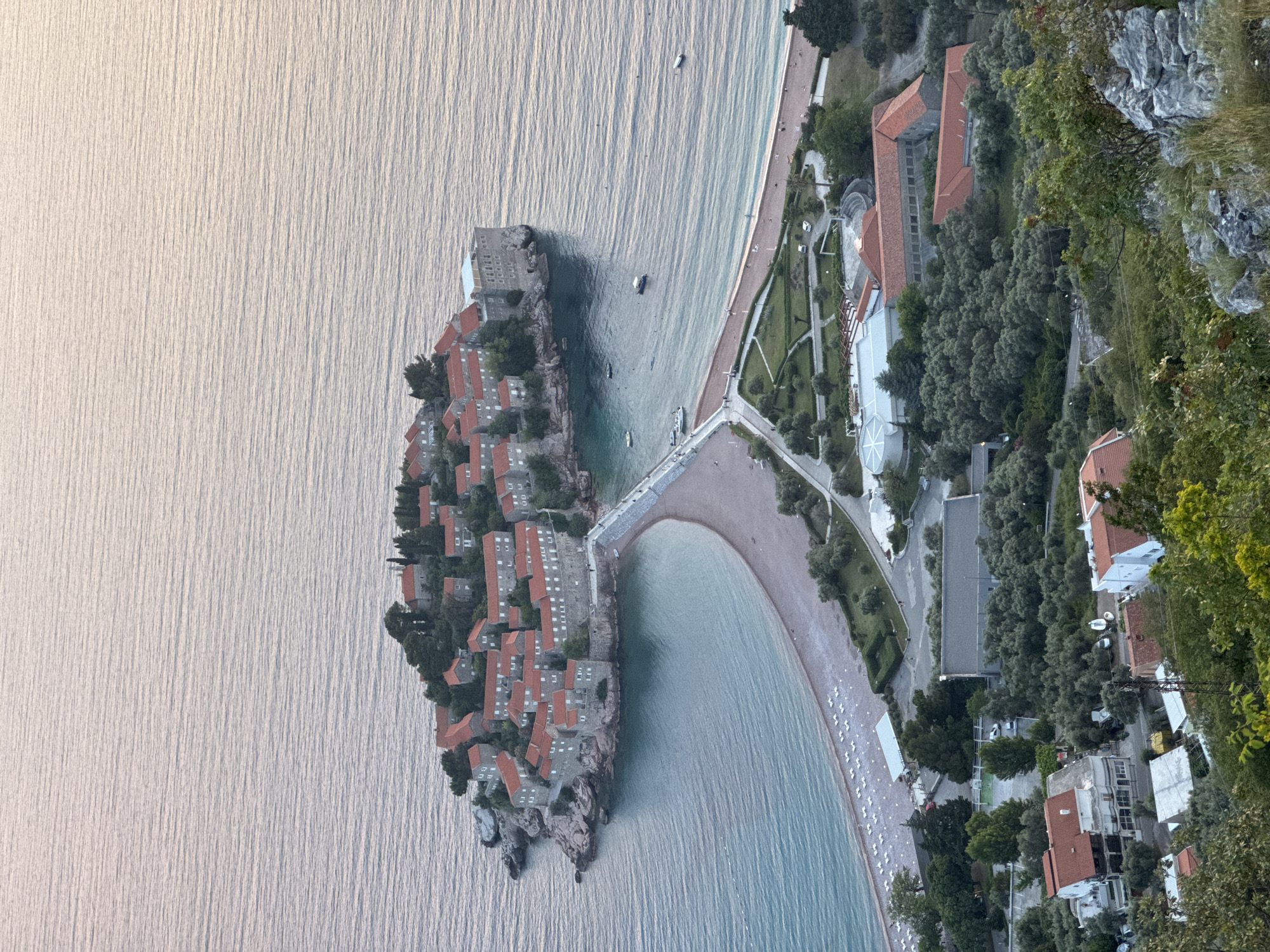 The image depicts the island of Sveti Stefan in Montenegro from an elevated perspective. The island is connected to the mainland by a narrow isthmus. The island is densely populated with buildings, most of which have red-tiled roofs. The water surrounding the island is calm. On the mainland side of the isthmus, there is a sandy beach with a line of white lounge chairs. Behind the beach is a strip of land with trees, gardens, and buildings, including some with red-tiled roofs. Farther inland, the land rises sharply and is covered with trees and some exposed rock formations. Some residential buildings are visible amongst the trees.