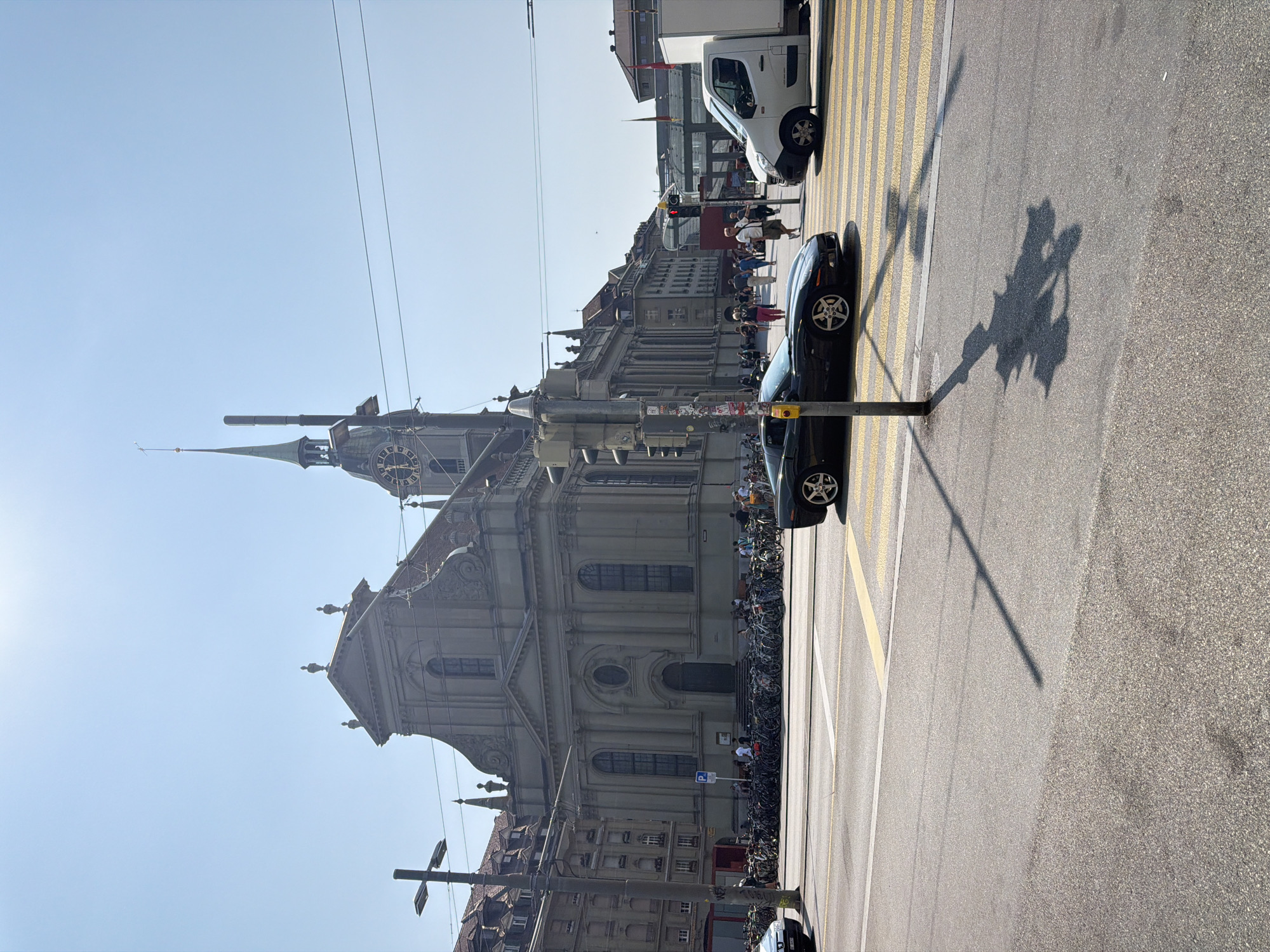 The image depicts a street scene in Bern, Switzerland, with a prominent church building in the background. The church, likely the Bern Minster, features a large clock tower with a spire. In the foreground, a black sports car is stopped at a pedestrian crossing. To the right, a white van is also at the crossing. Several people are visible walking on the sidewalks and crossing the street. A large number of parked bicycles are lined up in front of the church. The sky is clear and blue, indicating a sunny day. The street is paved with asphalt and marked with yellow and white lines for the pedestrian crossing and traffic lanes. Streetlights and power lines are also visible.
