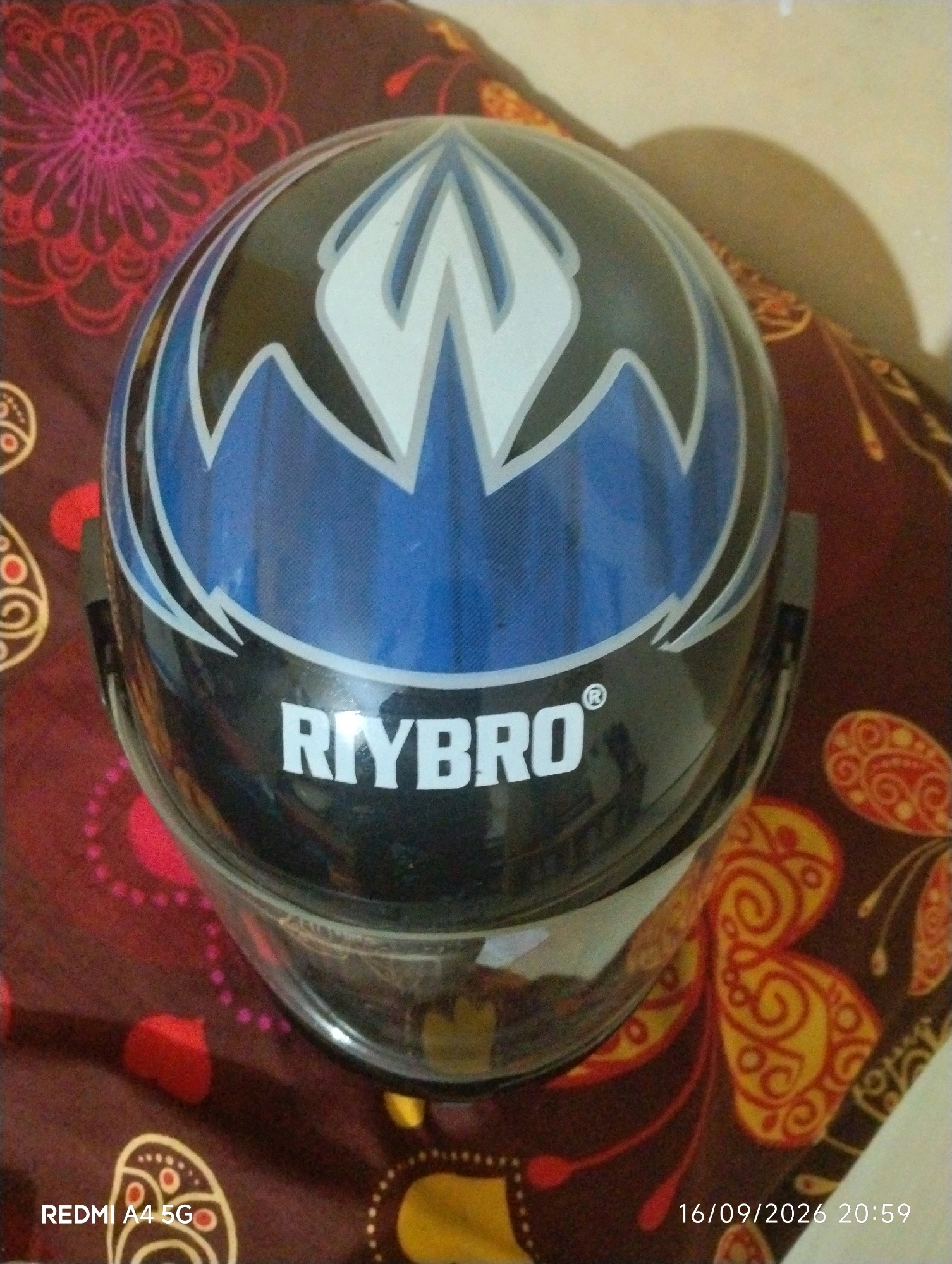 The image shows a close-up, eye-level view of a black and blue motorcycle helmet with white accents and the brand name "RIYBRO" printed on it in white capital letters. The helmet is resting on a patterned fabric surface, which appears to be a blanket or cushion, featuring red and orange floral and butterfly motifs on a dark brown background. A small red heart shape is visible on the fabric to the left of the helmet. The lighting suggests an indoor setting with a shadow cast on the fabric to the right of the helmet. The bottom left corner of the image displays the text "REDMI A4 5G", and the bottom right corner shows the date and time "16/09/2026 20:59". The location of Maluru, India, is not discernible from the visual information present in the image.