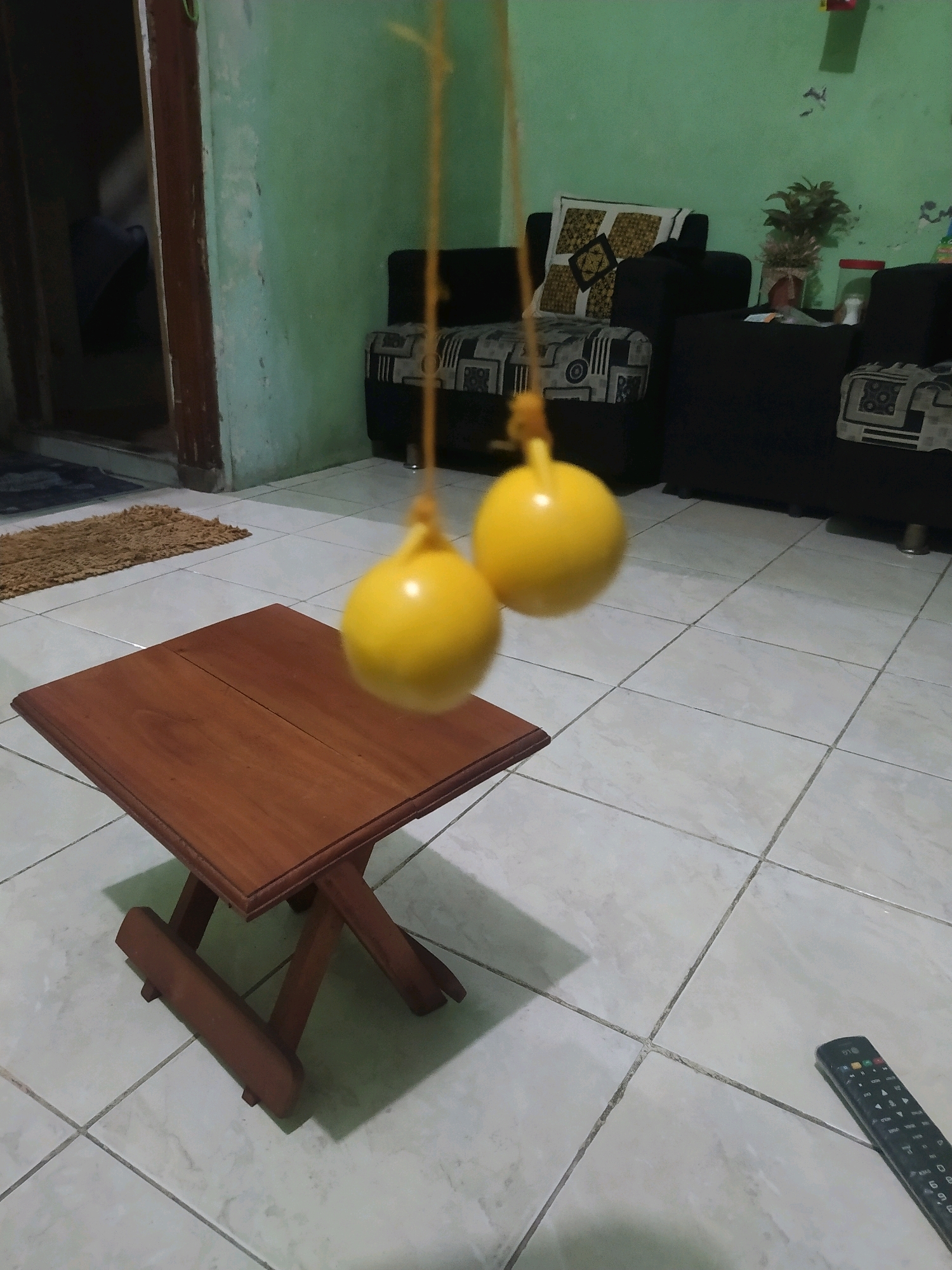 A low-angle, eye-level indoor shot shows a small, collapsible wooden table on a tiled floor. The table is in the foreground, with two bright yellow spheres, possibly children's toys, suspended from yellow strings above it. In the background, two dark sofas are visible against a light green wall. A potted plant sits on one of the sofas. To the right of the frame, a black LG television remote control lies on the floor. The scene is consistent with a residential interior in Sroyo, Indonesia.