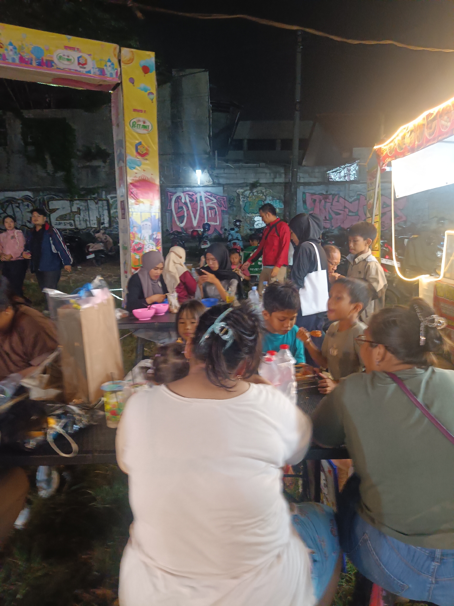 The image depicts an outdoor nighttime scene, likely at a local market or fair in Depok, Indonesia. Several people are gathered around tables, suggesting a social or casual dining event. The atmosphere appears lively, with a mix of adults and children engaged in various activities.
In the foreground, the backs of several individuals are visible, including a woman in a white top and another in a green top. They are seated or standing near a table laden with food and drinks. A young boy in a blue shirt is visible, holding a piece of food. Further back, a group of people are seated at a table, with two women wearing hijabs and a child. One woman is holding a pink bowl, and another is looking at her phone. A man in a red shirt stands behind them, facing away from the camera. Several other individuals are scattered throughout the scene, some standing and observing.
The setting includes food stalls or temporary structures adorned with colorful banners featuring cartoonish castles and balloons. Graffiti is visible on a wall in the background, adding to the urban, potentially informal, atmosphere. The lighting is predominantly artificial, with bright lights from the stalls casting a warm glow on the scene, indicating it is nighttime. The presence of motorcycles parked in the background suggests a local transportation method. The overall mood seems relaxed and convivial, with people enjoying food and each other's company. There is no discernible text that is clearly legible beyond what is on the banners.