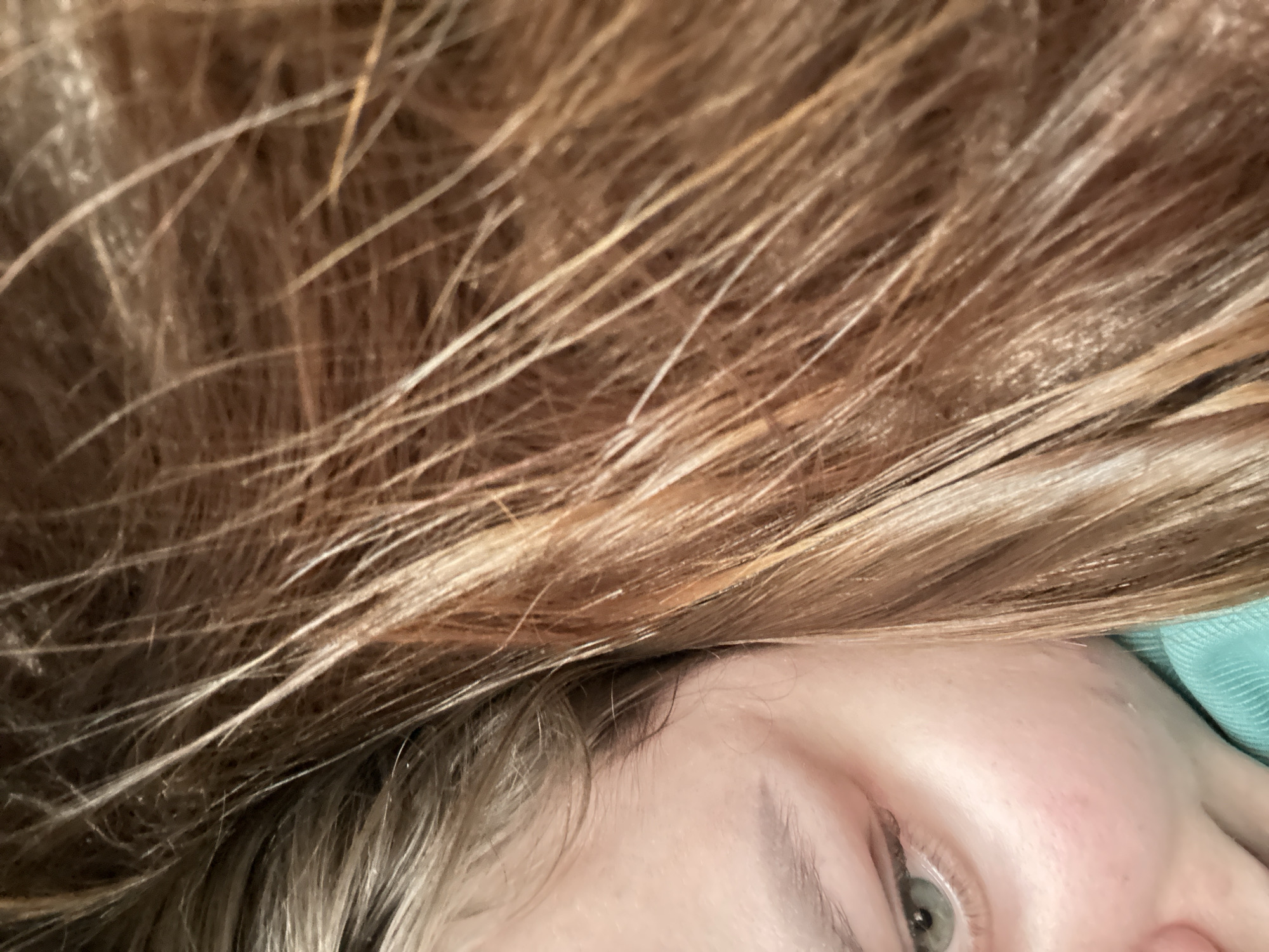 A close-up photograph shows a person's face partially obscured by their hair. The hair is a mix of blonde and light brown shades. An eye with a greenish iris is visible. Part of the person's cheek and eyebrow are also visible. The background is out of focus and includes a glimpse of what appears to be a light blue fabric. There is no information to suggest the image was taken in Chaikovsky, Russia.