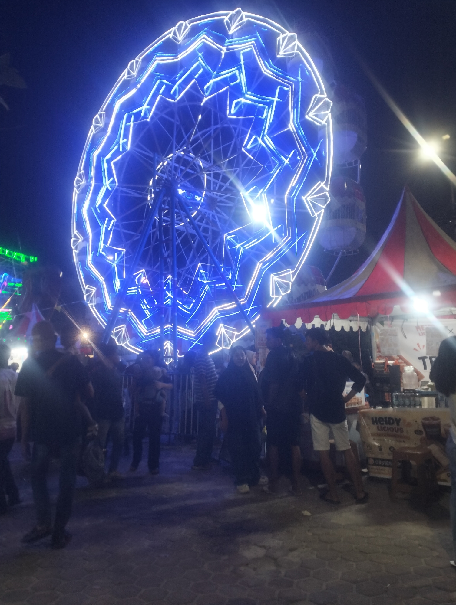 The image depicts a nighttime scene at what appears to be a carnival or fairground in Dumai City, Indonesia. The dominant feature is a large Ferris wheel, illuminated with vibrant blue and white lights, forming intricate patterns. In the foreground, a group of people are gathered, some standing and observing the Ferris wheel, while others are closer to a stall or booth. The stall has a red and white striped awning and displays a sign that reads "HEIDY LICIOUS". The ground is paved with hexagonal bricks. The overall atmosphere suggests a lively public event.