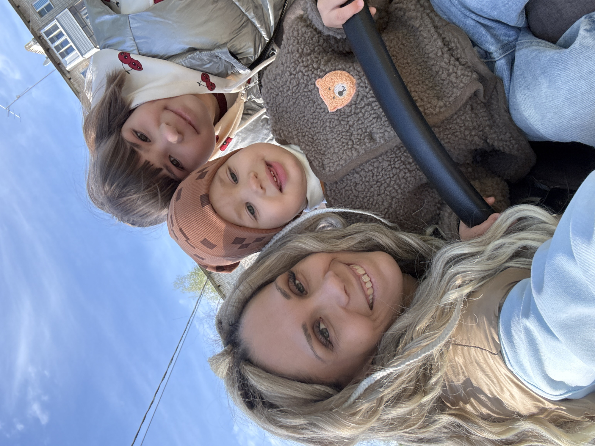 Three individuals, likely family members, are captured in a close-up, low-angle selfie. The person in the foreground, presumably an adult female, has long, blonde hair and wears a light blue hoodie and a beige vest. To her upper left is a young child wearing a brown hat. Above the child is another female, seemingly an older child, with dark hair and a sweatshirt with a cherry pattern. A brown jacket with a bear patch is visible near the younger child. In the upper background, the sky is visible, with a building at the upper left. Overhead electric wires pass across the frame. The location is identified as Rusanovo, Russia. The subjects are smiling, suggesting a positive interaction. The photo is taken in daylight.