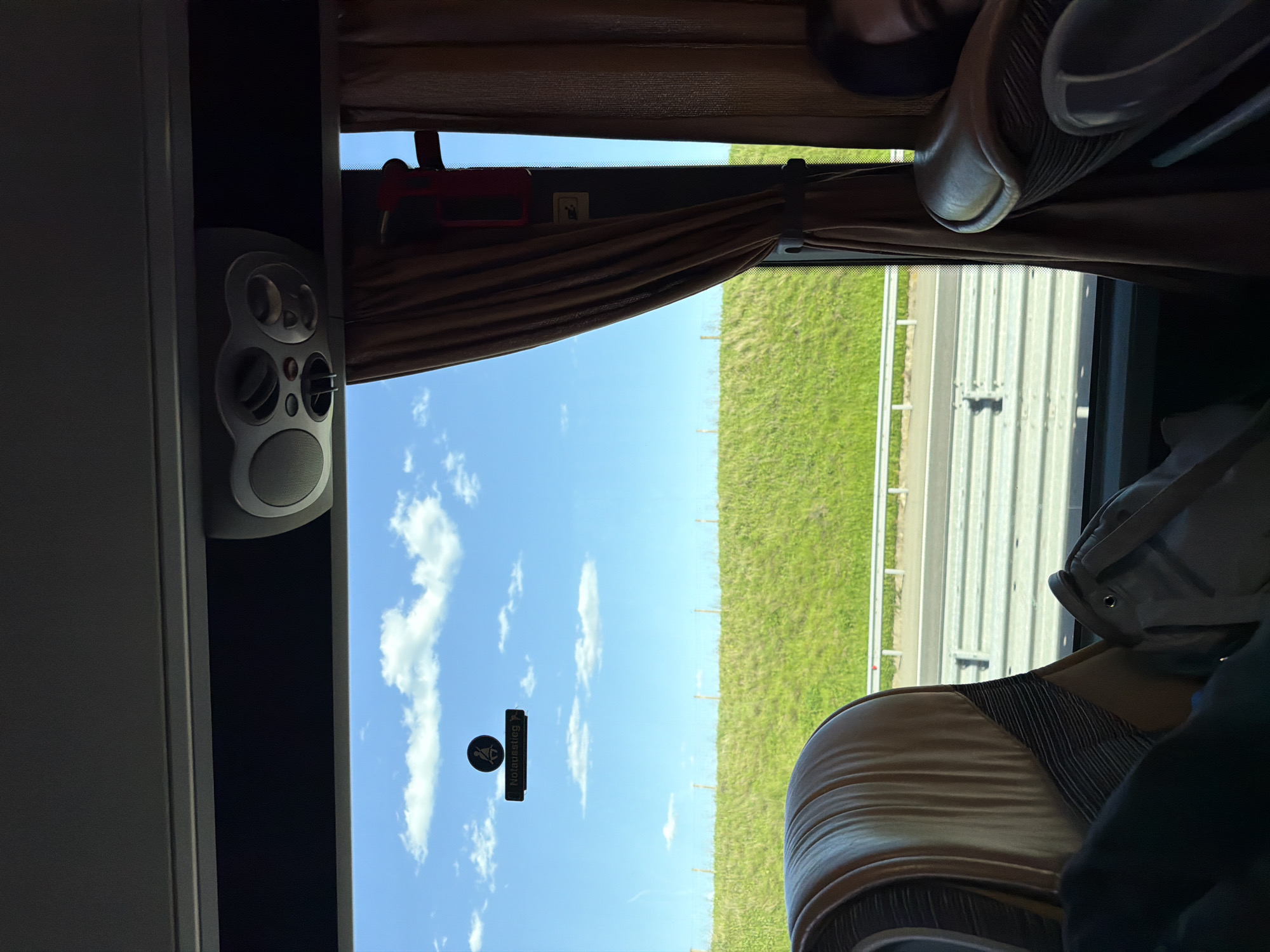 The image appears to be taken from inside a moving vehicle, potentially a bus or coach. The window view includes a blue sky with scattered clouds, a green grassy area, and a highway with a guardrail. Inside the vehicle, there is a speaker system mounted on a panel, brown curtains partially drawn, and tan upholstered seats. A sign written in German, possibly indicating an emergency exit, is visible. This does not provide enough information to place this photo in Malko Tranovo, Bulgaria.