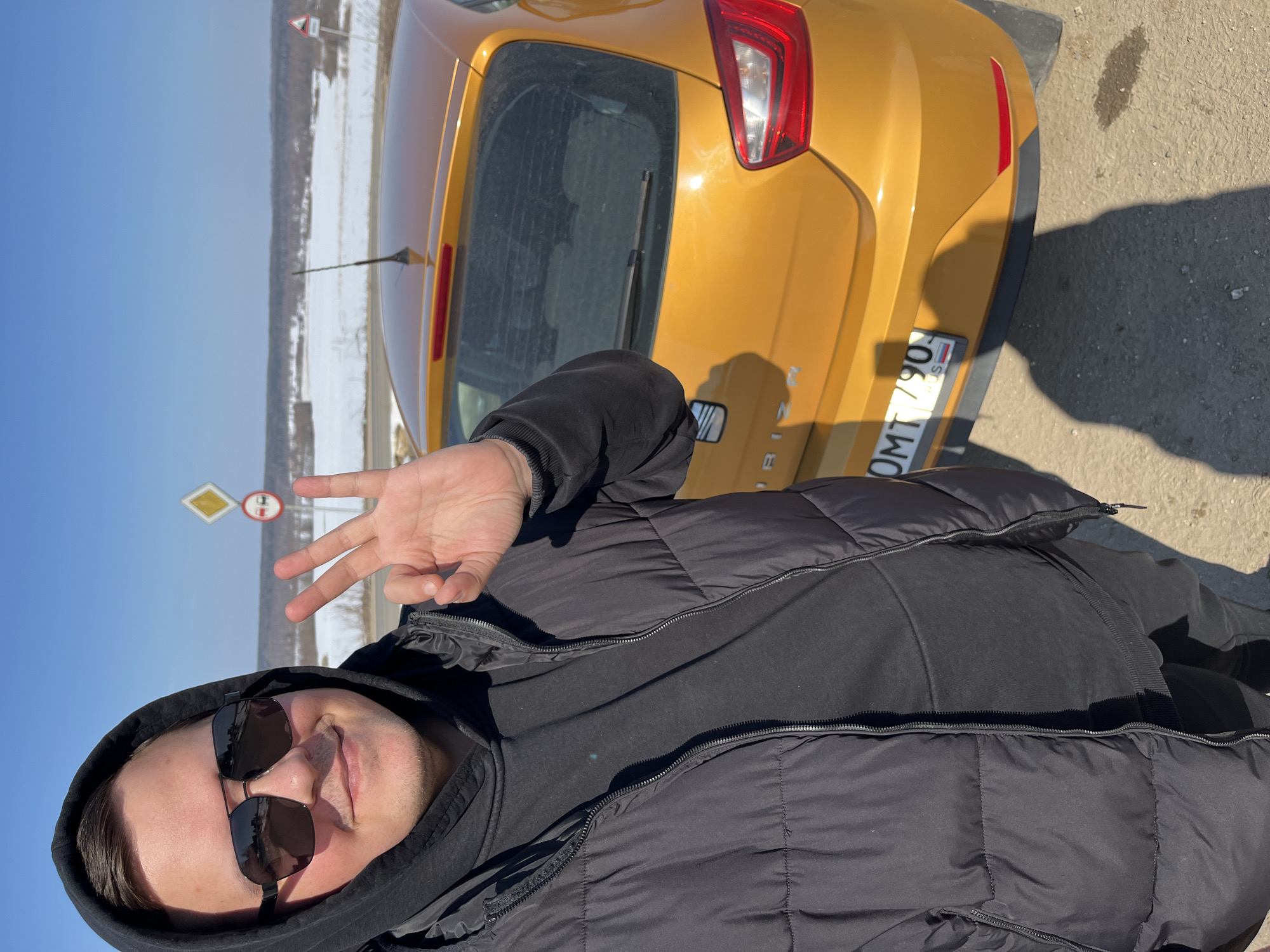 A solitary male subject is depicted in a selfie, taken outdoors in Zubovo, Russia, under clear blue skies. The subject, exhibiting a slight smile, wears dark aviator-style sunglasses, a black hooded sweatshirt, and an unzipped black puffer jacket. His right hand is raised, forming an "OK" gesture with thumb and forefinger touching. Positioned behind and to the right of the subject is the rear of a bright orange SEAT Ibiza hatchback, bearing a Russian license plate with characters "OMT 790". The vehicle's rear window appears dusty. In the background, a paved road runs alongside a snow-covered plain with sparse, bare deciduous vegetation, extending to a distant treeline. Two prominent road signs are visible: a yellow diamond-shaped 'main road' sign and a white circular 'no overtaking' sign with a red border and two vehicle silhouettes. Bright, direct sunlight casts a distinct shadow of the subject onto the asphalt surface in the foreground. The overall scene suggests late winter or early spring conditions.