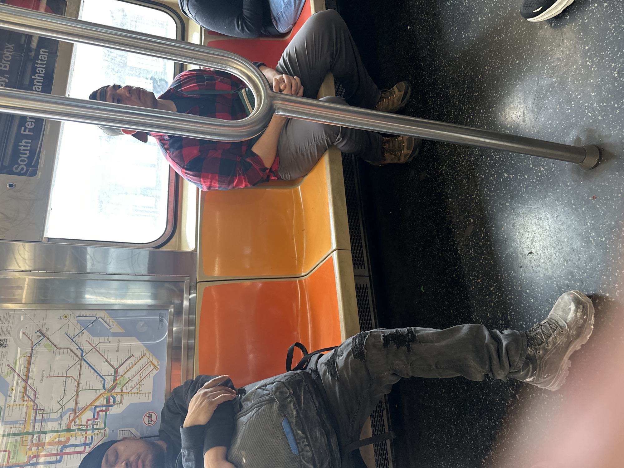 The image shows the interior of a New York City subway car. Two people are visible. One person is seated and wearing a red and black flannel shirt, gray pants, and brown hiking boots. They are holding onto the subway pole. Another person is lying down across the seats, wearing a black hooded sweatshirt, gray pants, and gray boots. A subway map is visible on the wall of the car. The subway car is orange. The subway car has a sign that says “South Fer”.