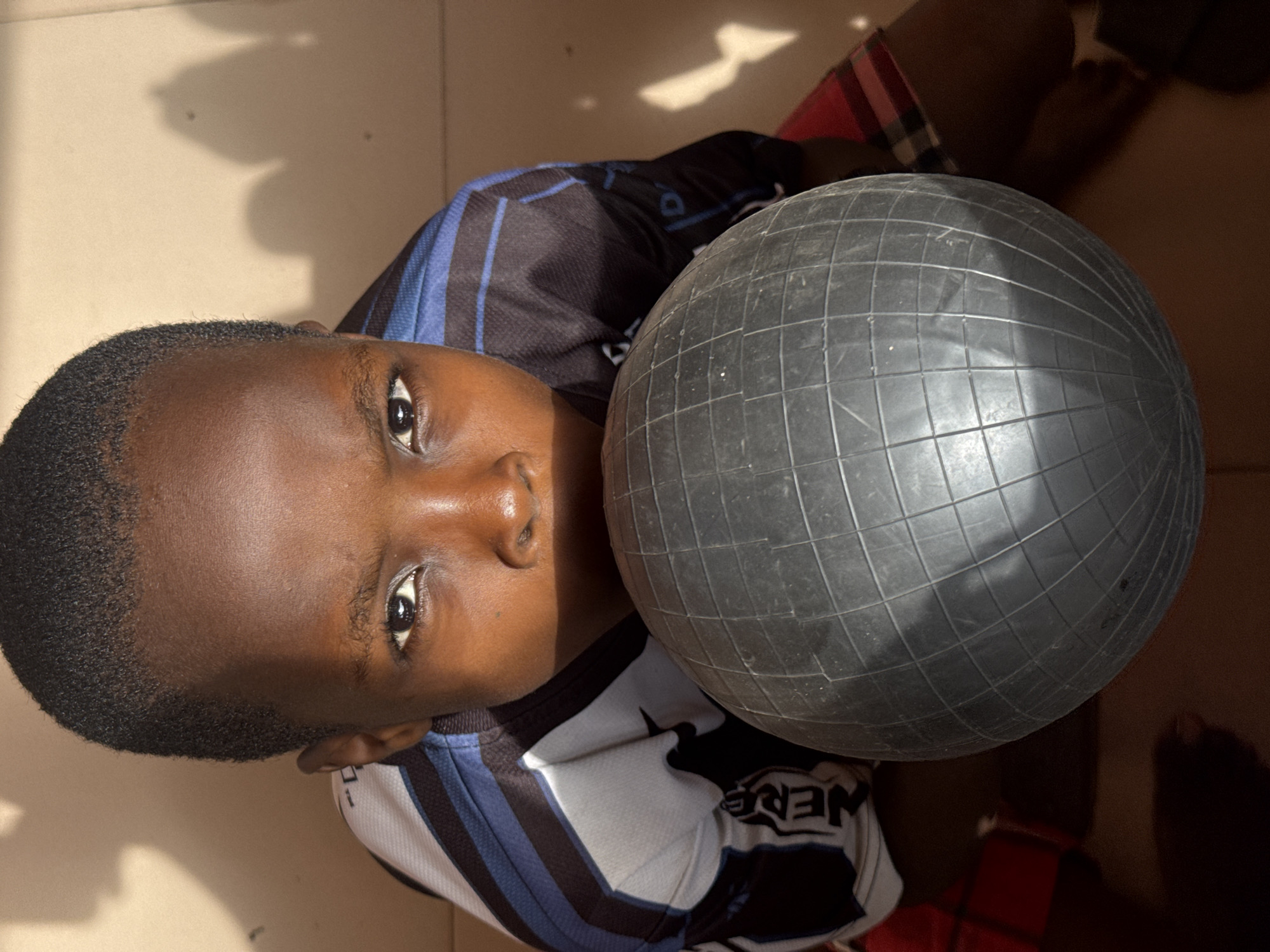 A young, dark-skinned child is holding a dark gray, grid-patterned ball. The child is wearing a sports jersey with blue and white stripes. The location appears to be indoors in the city of Onitsha, Nigeria, with a tile or concrete floor visible. The child is looking upwards. The lighting is partially sunny, with shadows cast across the scene. Part of the child's legs and feet are visible.