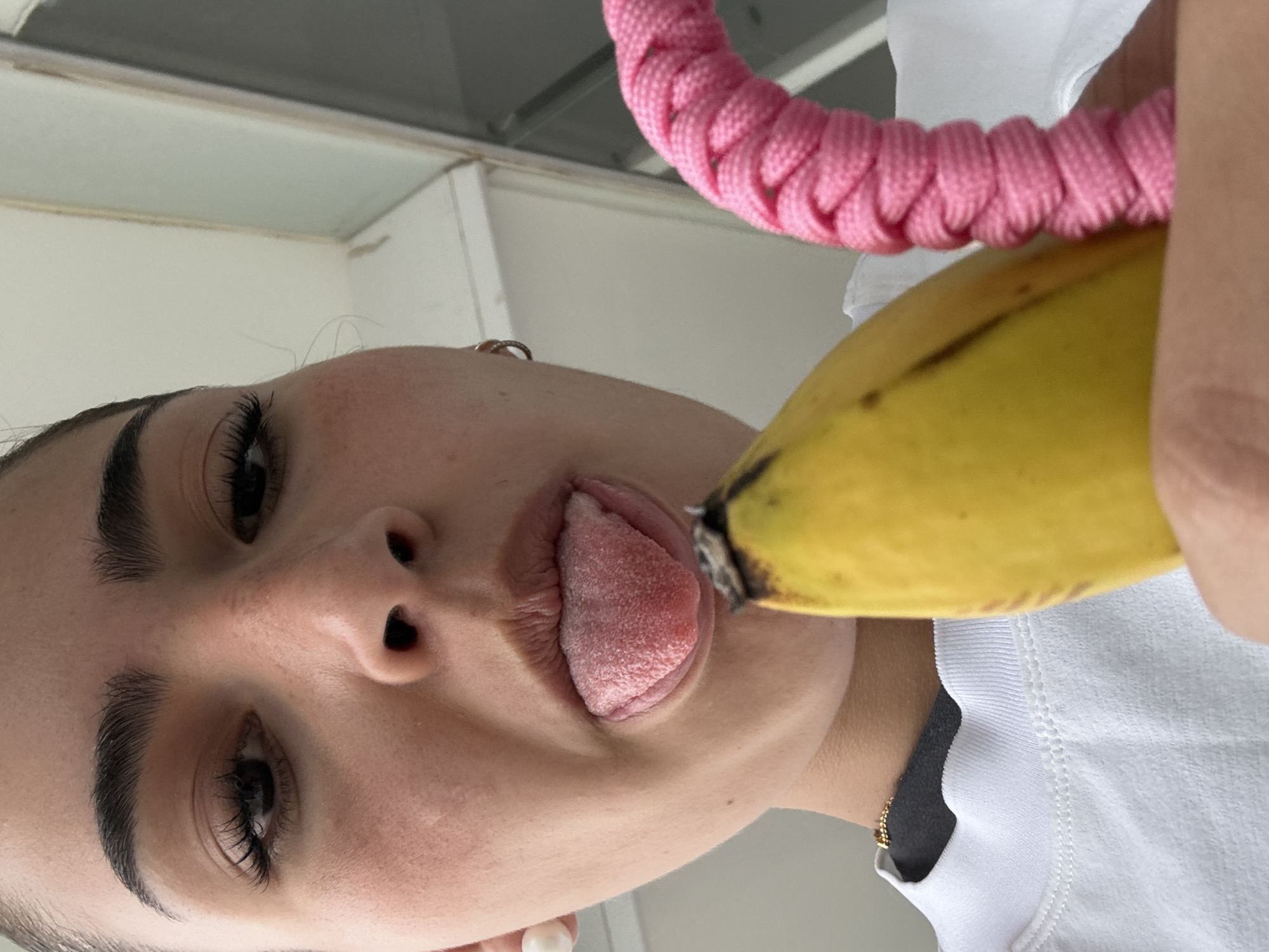 A person is holding a banana to their mouth, extending their tongue to touch it. The banana is mostly yellow with some brown near the end. A pink coiled object is also visible. The person is wearing a white t-shirt. The photograph is taken from a low angle, looking up at the subject.
The location is unknown.