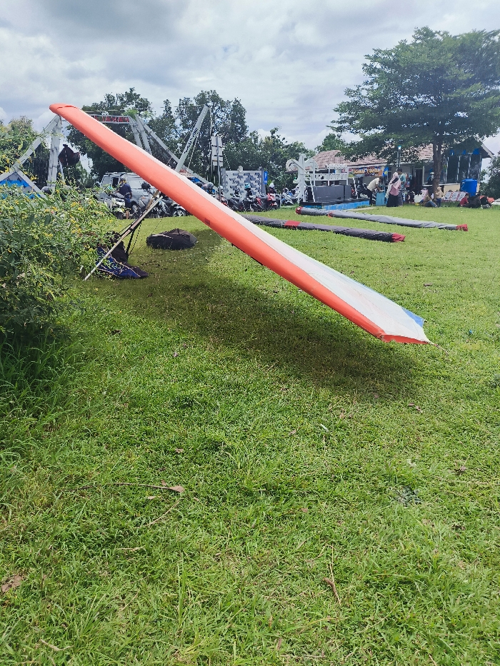 The image shows a red and white hang glider lying on a grassy field. The hang glider is lying on its side, with the wing extended and the frame on the ground. It appears to have been used recently. There is a small amount of debris scattered nearby. The location is a park in the city of Majalengka, Indonesia, at the coordinates provided. There are no people in the image. The weather is clear and the sky is partly cloudy. The grass is green and lush, the ground is level, and the surrounding area appears to be a park or recreational area. The hang glider is lying in the middle of the image, and there is a tree in the background. There are also some buildings visible in the distance. There are two other hang gliders lying on the grass behind the subject, one is red and white, the other is blue and white. The location is consistent with a park or recreational area where people might go for hang gliding.