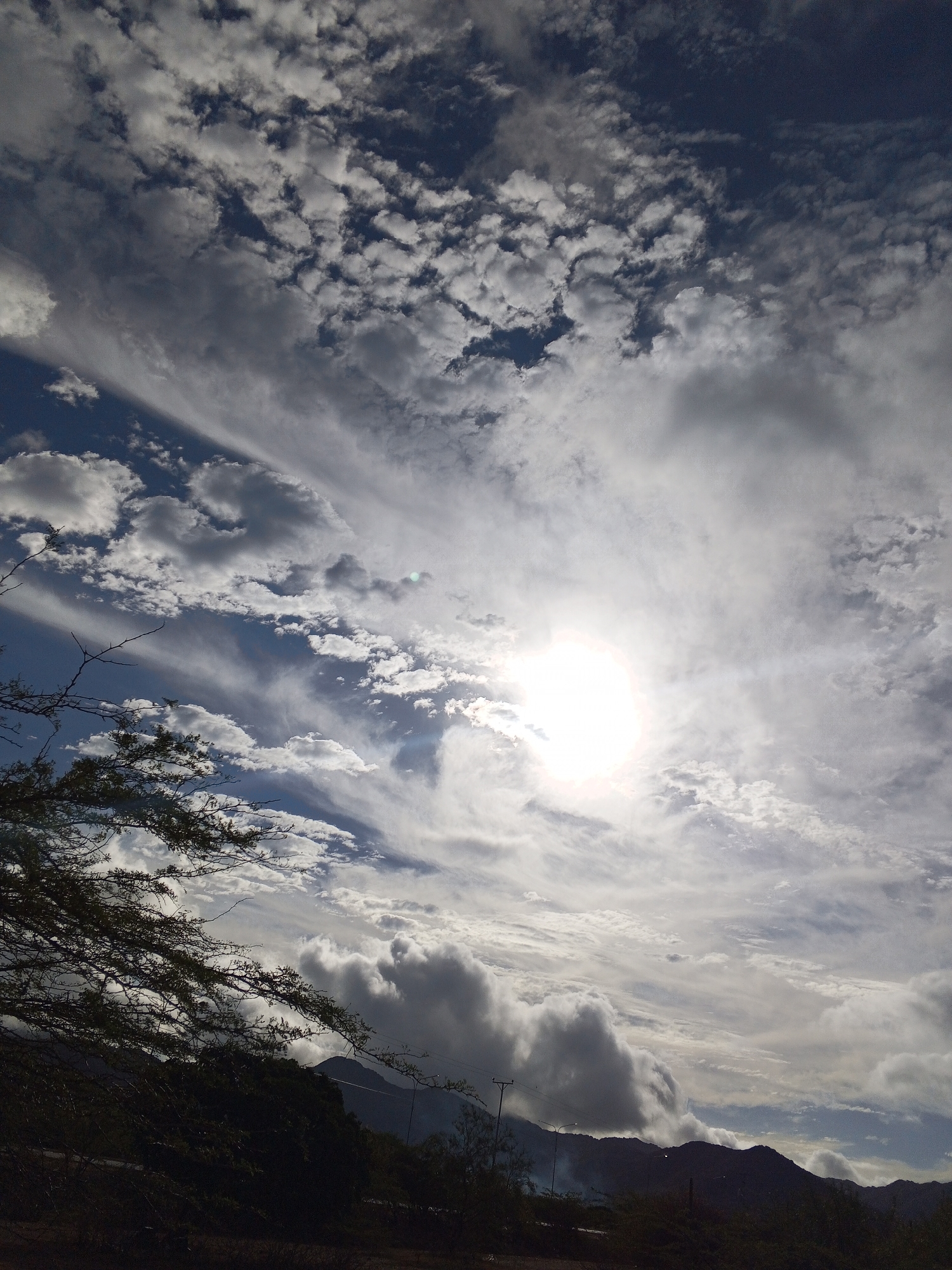 The image depicts a cloudy sky over a mountainous landscape, potentially in Altagracia, Venezuela. The sun is visible through a break in the clouds, creating a bright spot. The sky contains a mix of cumulus and cirrus clouds. In the lower portion of the image, silhouettes of trees and mountains are visible. There is some evidence of light pollution.