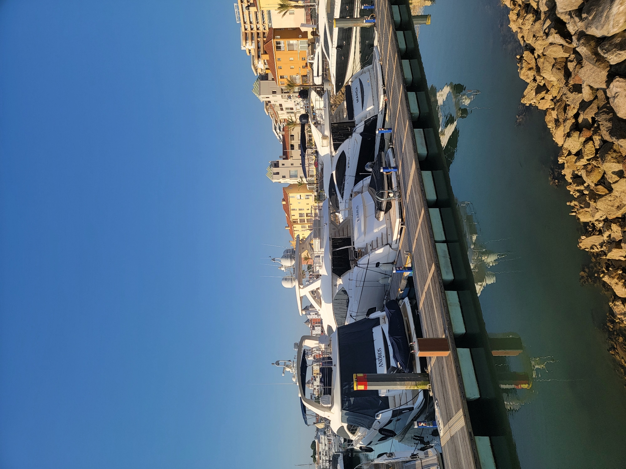 A waterfront scene in Quarteira, Portugal, features several luxury yachts docked side-by-side at a wooden pier under a clear blue sky. The yachts are white with dark accents and appear to be various models and sizes. One yacht in the foreground has "ANDROS" written on its stern. The pier is constructed of wooden planks supported by green floats. Buildings with light yellow and orange facades, characteristic of Mediterranean architecture, are visible in the background, rising up a gentle slope. Some buildings have distinctive green tiled roofs. Palm trees are interspersed among the buildings. The water is a deep green, reflecting the sky and the yachts. The foreground is bordered by a rocky shoreline. No people are visible in the image.
