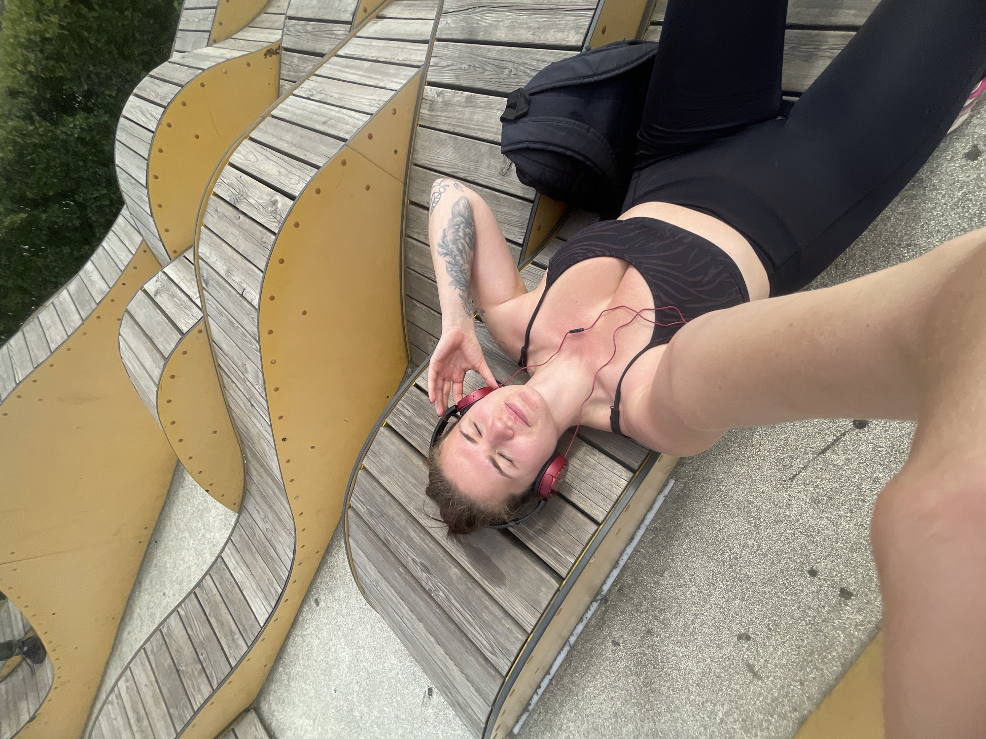 A woman is lying on a curved wooden structure, possibly a bench or seating area. She is wearing a black sports bra and black leggings. She has headphones on and appears relaxed, with her eyes closed. Next to her is a black backpack. The structure she is lying on is composed of multiple wooden planks, curved in a wave-like pattern, with yellow supports on the sides. The background includes greenery and concrete surfaces. The location appears to be an urban park or public space. The city is unknown.