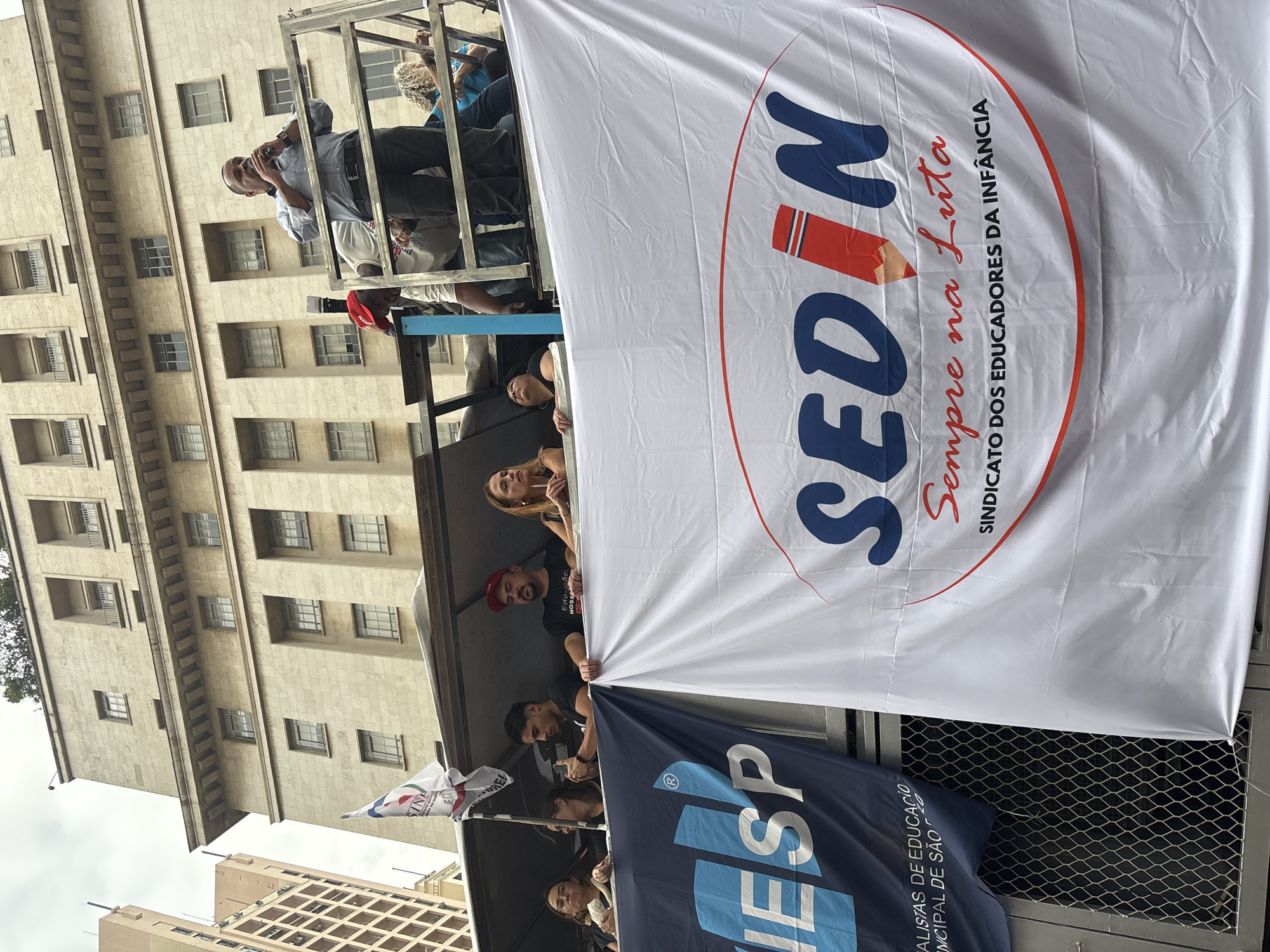 The image captures an outdoor demonstration or rally taking place in São Paulo, Brazil. Several individuals are visible on a truck or float, with large banners prominently displayed.
The most prominent object is a large white banner, featuring a blue "SEDIN" logo stylized with a red pencil. Below this, the red cursive text reads "Sempre na Luta" (Always in the Fight), followed by "SINDICATO DOS EDUCADORES DA INFANCIA" (Union of Early Childhood Educators) in smaller black letters. Below this, a dark blue banner is partially visible, showing light blue text segments like "ESP", "ALISTAS DE EDUCAÇÃO", and "NICIPAL DE SÃO", strongly suggesting a municipal education workers' union from São Paulo. A smaller white flag, partially visible, displays the words "Educão:" and "NOSSA", possibly part of a slogan.
Among the people on the platform, an older man with glasses is seen speaking into a microphone, appearing to address a crowd. Other individuals, including men and women, are holding the banners, with some looking towards the viewer. Their expressions are generally neutral or focused.
The background features tall, light-colored concrete buildings with multiple windows, characteristic of an urban environment. The sky above is overcast, suggesting a cloudy daytime event. The overall scene conveys a sense of collective action and advocacy, likely related to educators' rights and conditions.