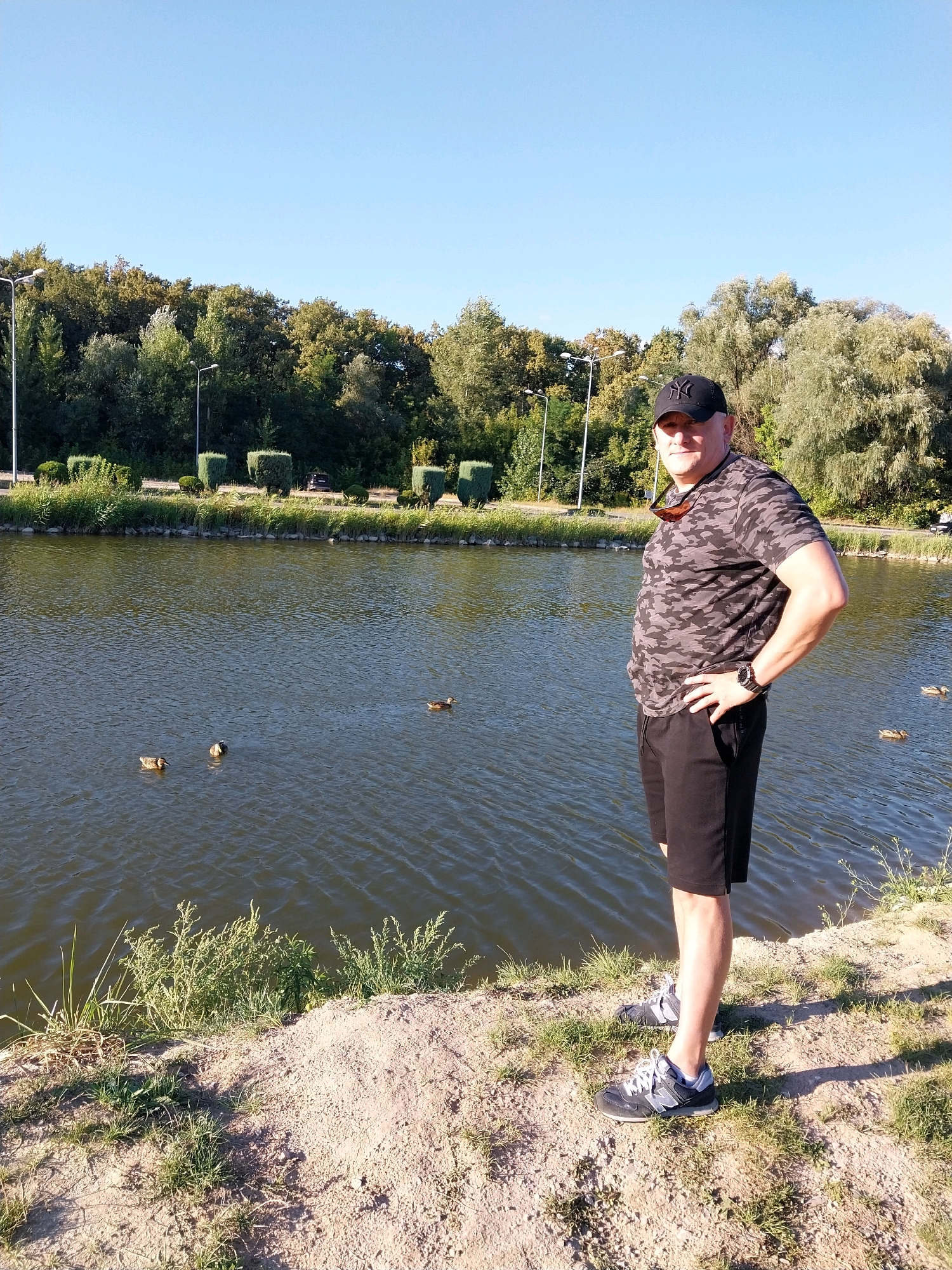 A man is standing on the shore of a lake, with a forested area and a clear blue sky in the background. The man is wearing a camouflage t-shirt, black shorts, a black baseball cap, and athletic shoes. He has his left hand on his hip and is looking at the camera. There are three ducks swimming in the lake in front of him. The shore is covered in sand and grass. The scene appears to be located in Dnipro, Ukraine.