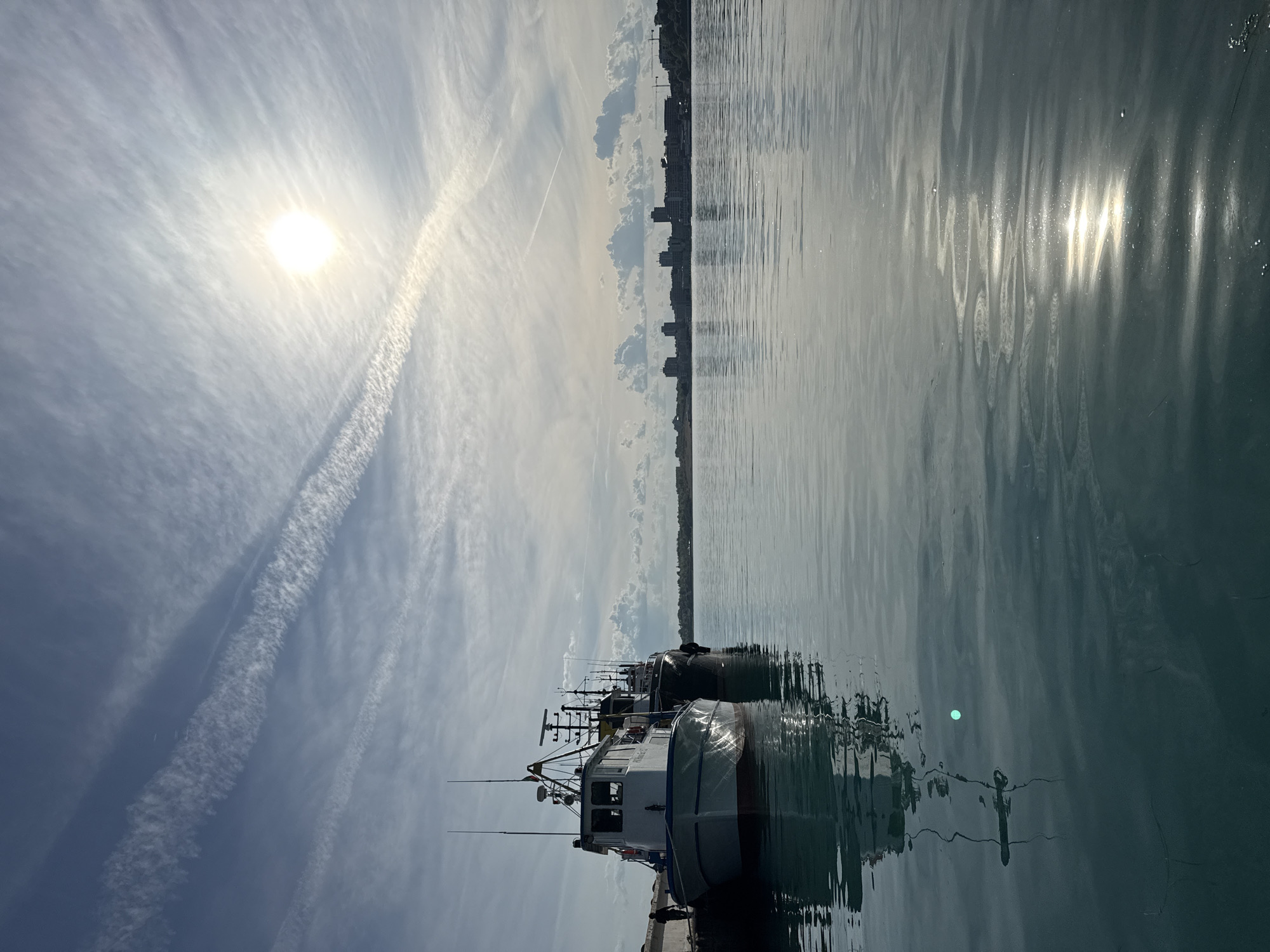 The image depicts a waterfront scene in Nesebar, Bulgaria. Several fishing boats are docked along a concrete pier. In the background, a cityscape is visible across the water. The sky is partly cloudy with prominent contrails from aircraft. The sun is visible and casting a bright reflection on the water's surface, which is rippled. The overall atmosphere is bright and sunny.