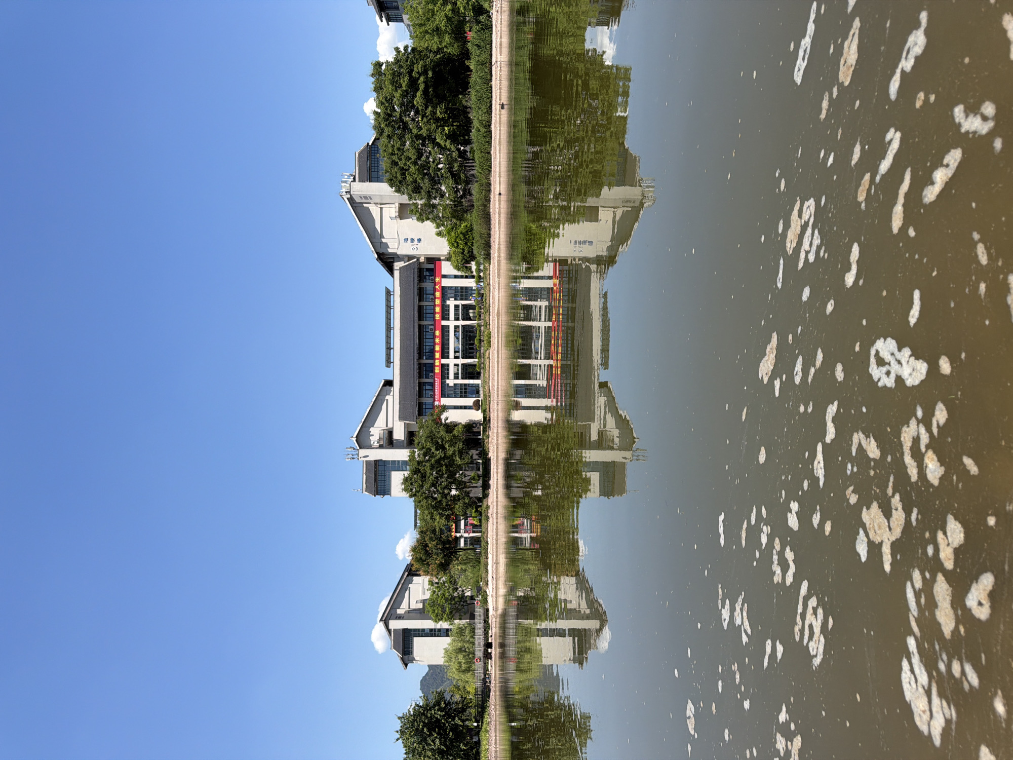 In the city of Jiangjin, China, a serene lake reflects a modern building complex under a clear blue sky. The water's surface is disturbed by a scattering of light-colored foam, creating an abstract pattern in the foreground. The building, with its sharp angles and numerous windows, is partially obscured by lush green trees lining the shore. A footbridge extends from the bank towards the building, adding a touch of human infrastructure to the natural landscape. The reflection of the building and trees on the calm water creates a symmetrical and visually appealing composition. The overall scene depicts a tranquil, sun-drenched day in a well-maintained urban environment.