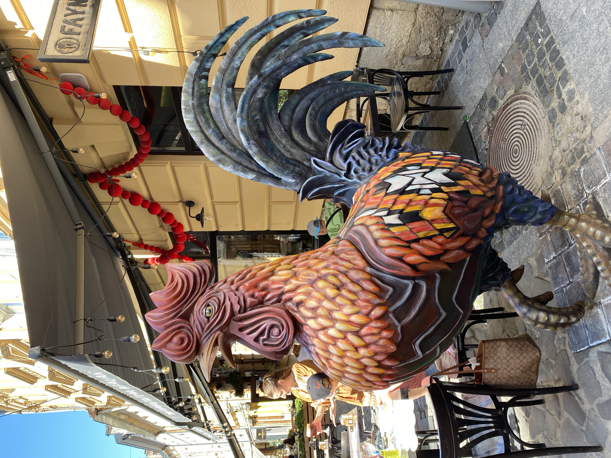 The image depicts a large, colorful sculpture of a rooster situated on a cobblestone street in Lviv, Ukraine. The rooster sculpture is the primary subject, with its body adorned in vibrant hues of red, orange, yellow, and blue. Its tail feathers are a deep blue with iridescent green accents.
Behind the rooster, a building with a yellow facade is visible, featuring awnings and outdoor seating for a cafe or restaurant. A sign reading "FAYNA" with a fork and knife icon is mounted on the building. Red, velvet-like spherical decorations are strung along the awnings, adding a festive touch.
In the mid-ground, a woman in a brown dress is standing near a table, holding a drink and looking towards the rooster. A young child wearing a blue cap is seated at another table. Several outdoor tables and chairs are arranged on the cobblestone pavement. A brown designer handbag hangs from the back of one of the chairs.
The overall scene suggests a lively urban setting, likely a pedestrian street in Lviv, with the rooster sculpture serving as a prominent decorative element for a local establishment. The lighting indicates it is daytime, with clear skies and bright sunshine casting shadows.