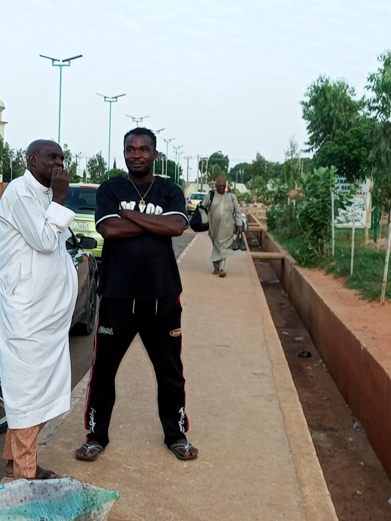 In Gombe, Nigeria, two men stand on a sidewalk next to a street. The man on the left wears a white robe and is seen from the chest down, his hand near his mouth. The man on the right wears a black t-shirt with "NEW YORK" printed on it and black track pants. He has his arms crossed and is wearing a necklace. In the background, a third man in a light-colored robe walks away from the camera, carrying two black bags and a backpack. A green vehicle is parked behind the first two men. The sidewalk runs alongside a drainage ditch filled with water and debris. Trees and streetlights line the street. A sign with text is visible in the distance. The sky is overcast.