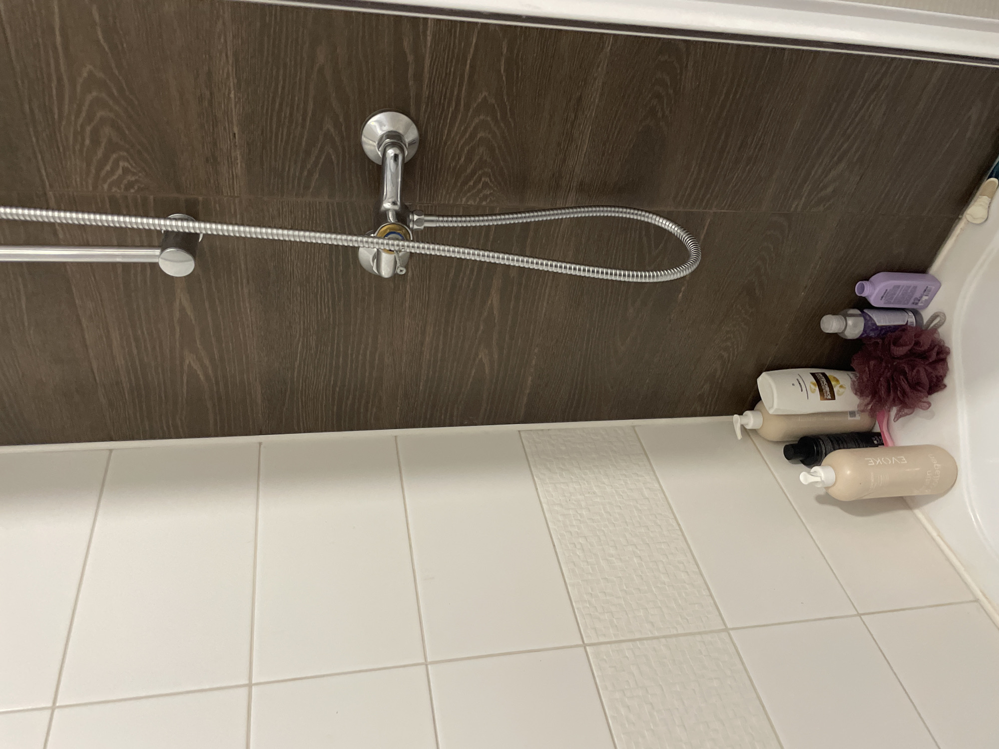 The image depicts a close-up view of a shower enclosure within a bathroom. The enclosure features white tiled walls on the left and a dark, wood-grained tiled wall on the right. A chrome shower fixture with a flexible hose is mounted on the wood-grained wall. Several bottles of toiletries, including shampoo and conditioner, are placed on the edge of the bathtub. Also visible is a pink mesh shower pouf. The scene is domestic and appears to be in a bathroom. The location is specified as Sinyavino, Russia.