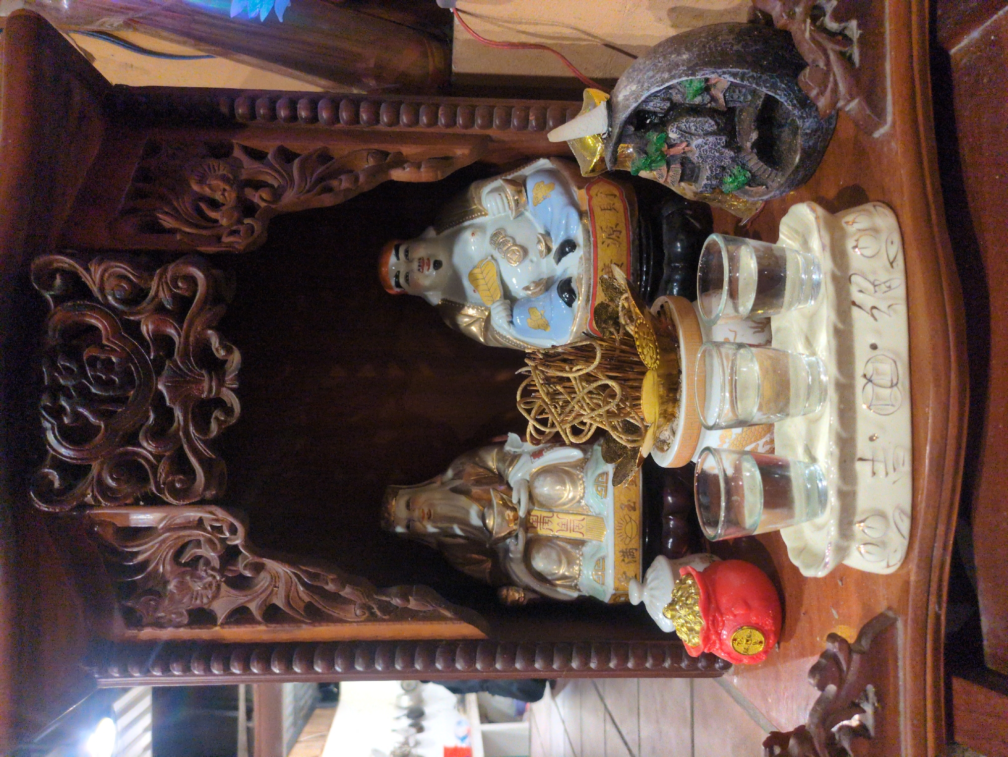 A tabletop shrine sits inside a wooden cabinet. The shrine includes two porcelain figurines, a small fountain, three glasses of liquid, and a golden decoration. A red bag rests at the bottom. The shrine is located in Phú Quốc, Vietnam.