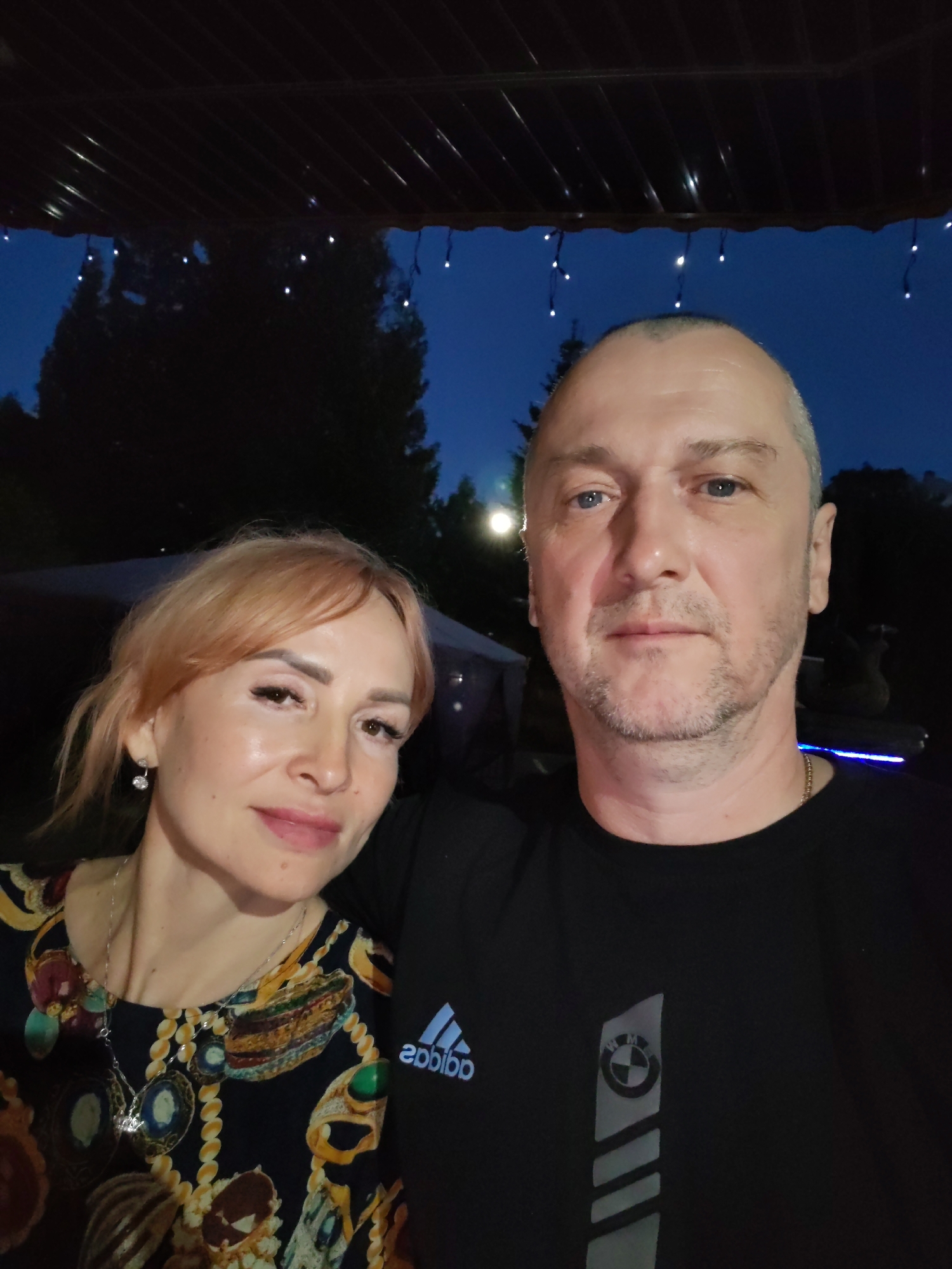 An outdoor selfie captures a middle-aged Caucasian man and woman posing for the camera. The man, with short grey hair and stubble, wears a black t-shirt with a BMW logo. The woman, with blonde hair and light makeup, sports a colorful patterned dress and a silver necklace. They are positioned closely together, suggesting a personal relationship. Behind them, the background is dark, with blurred trees and faint decorative lights, indicating it is likely evening or night. The scene appears to be in a relaxed, possibly recreational setting, such as a backyard or patio area.