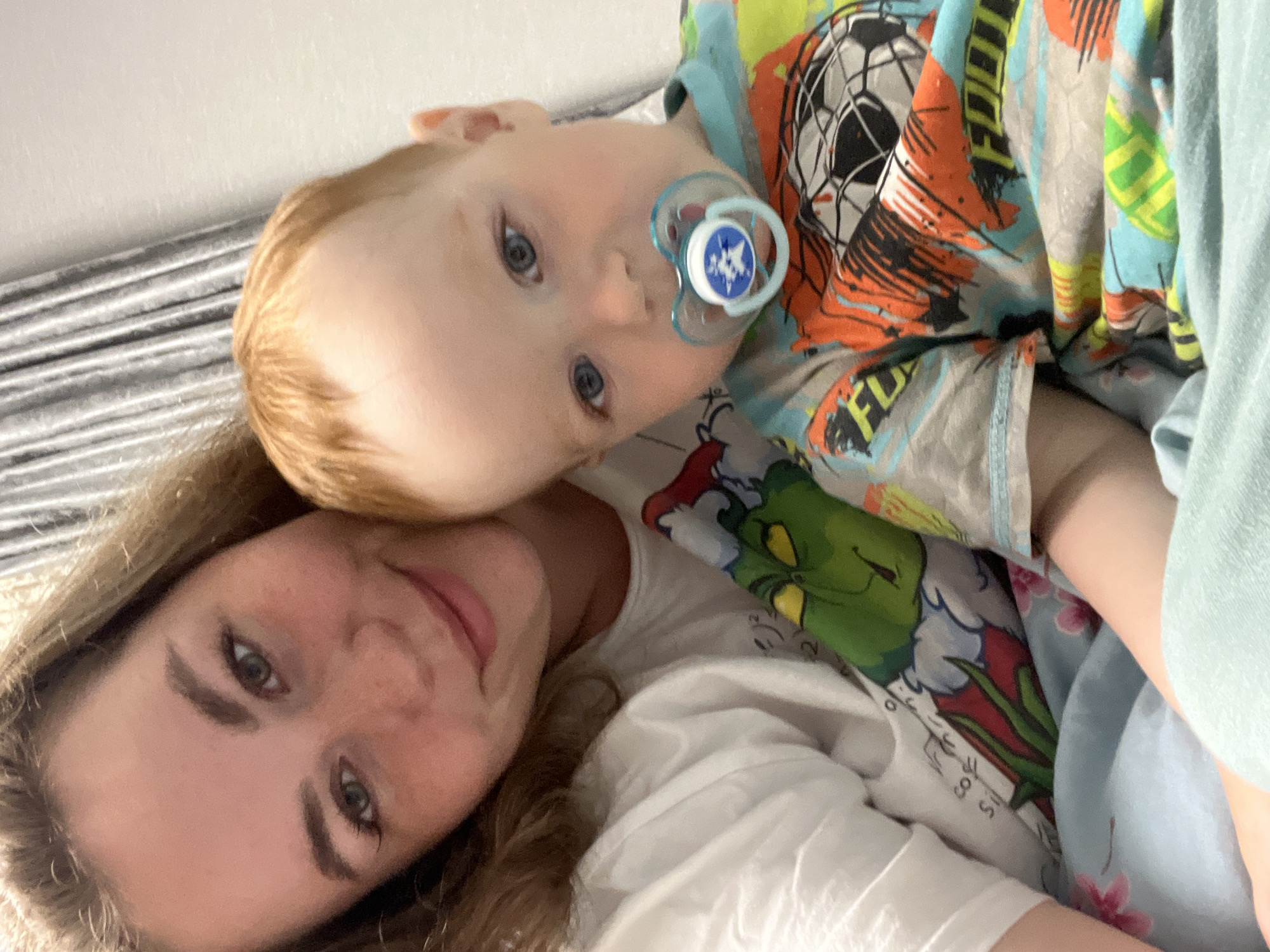 A photograph depicts a woman and a young child indoors. The woman has light skin, brown hair, and is lying on her side. The child, who has fair skin and reddish hair, is positioned above her with a pacifier in their mouth. The child is wearing a patterned shirt. Beneath them is a white garment featuring a green character. The image's location is Chaikovsky, Russia.