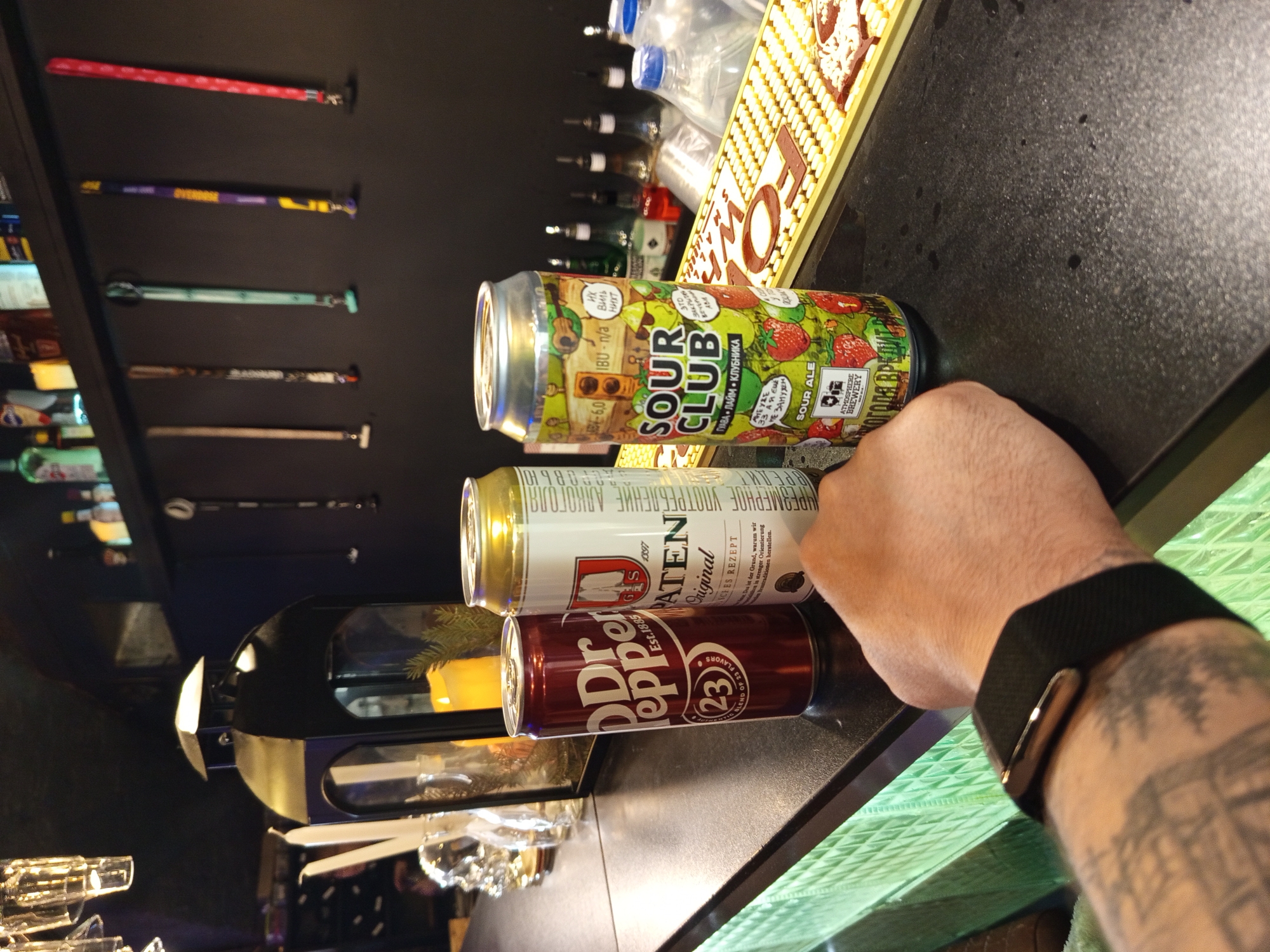 This is a close-up, vertical shot of a person's hand holding two cans of soda and a can of sour ale. The hand, which has tattoos, is wearing a black watch. In front of the cans, a portion of a black surface with a textured pattern is visible, with some condensation and small dark spots. To the right of this surface, a bright green, patterned surface can be seen.
The cans are stacked. The top can, a sour ale named "Sour Club," is colorful and features images of strawberries and musical instruments. Text on the can includes "Sour Ale," "Guava Lime Strawberry," and "Atmosphere Brewery." Below it is a white and gold can labeled "Alten Original." The bottom can is a deep red, a Dr. Pepper can, with "Est. 1885" visible.
In the background, a dark shelf is lined with various items, including what appear to be lanyards or straps with colorful designs. Further back, bottles are visible on another shelf, some with blue caps. The overall setting appears to be a bar or a similar establishment, possibly in Saint Petersburg, Russia, given the context. The lighting suggests it could be evening or indoors, as there are no clear indications of natural light. The focus is on the cans and the hand, suggesting the activity is related to enjoying these beverages.