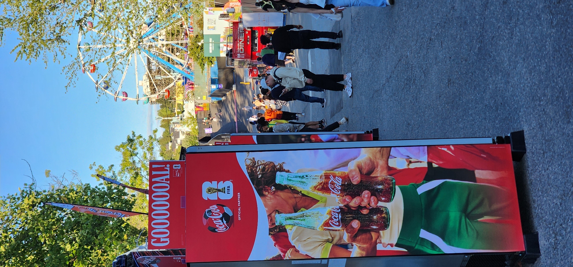 A tall, red Coca-Cola advertisement is displayed prominently on the left side of the frame. The advertisement features an image of two hands holding Coca-Cola bottles, and text that reads "GOOOOOOAL!" along with a FIFA World Cup logo. In the background, a Ferris wheel and various fairground attractions are visible, suggesting the scene takes place at a festival or amusement park. Several people are walking around in the background, some are holding phones, and others are observing the attractions. The overall setting is an outdoor area with trees and a clear blue sky.