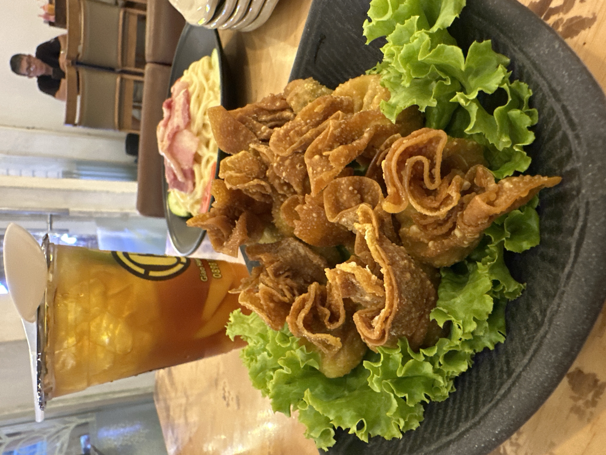 This is a close-up, angled shot of a plate of food at a restaurant. In the foreground, a dark grey plate is topped with a bed of green lettuce. On top of the lettuce are golden-brown, deep-fried wontons with a ruffled, flower-like appearance. To the left, a plastic cup filled with iced tea, complete with a lid and spoon, is visible. The cup has a logo that appears to read "Giao Hi...0898." In the background, a plate of noodles with some strips of meat on top sits next to the plate of wontons. Tables and chairs are visible in the soft background, and a person is seated at a table in the distance. The photo was taken in Bac Nha Trang Ward, Vietnam.