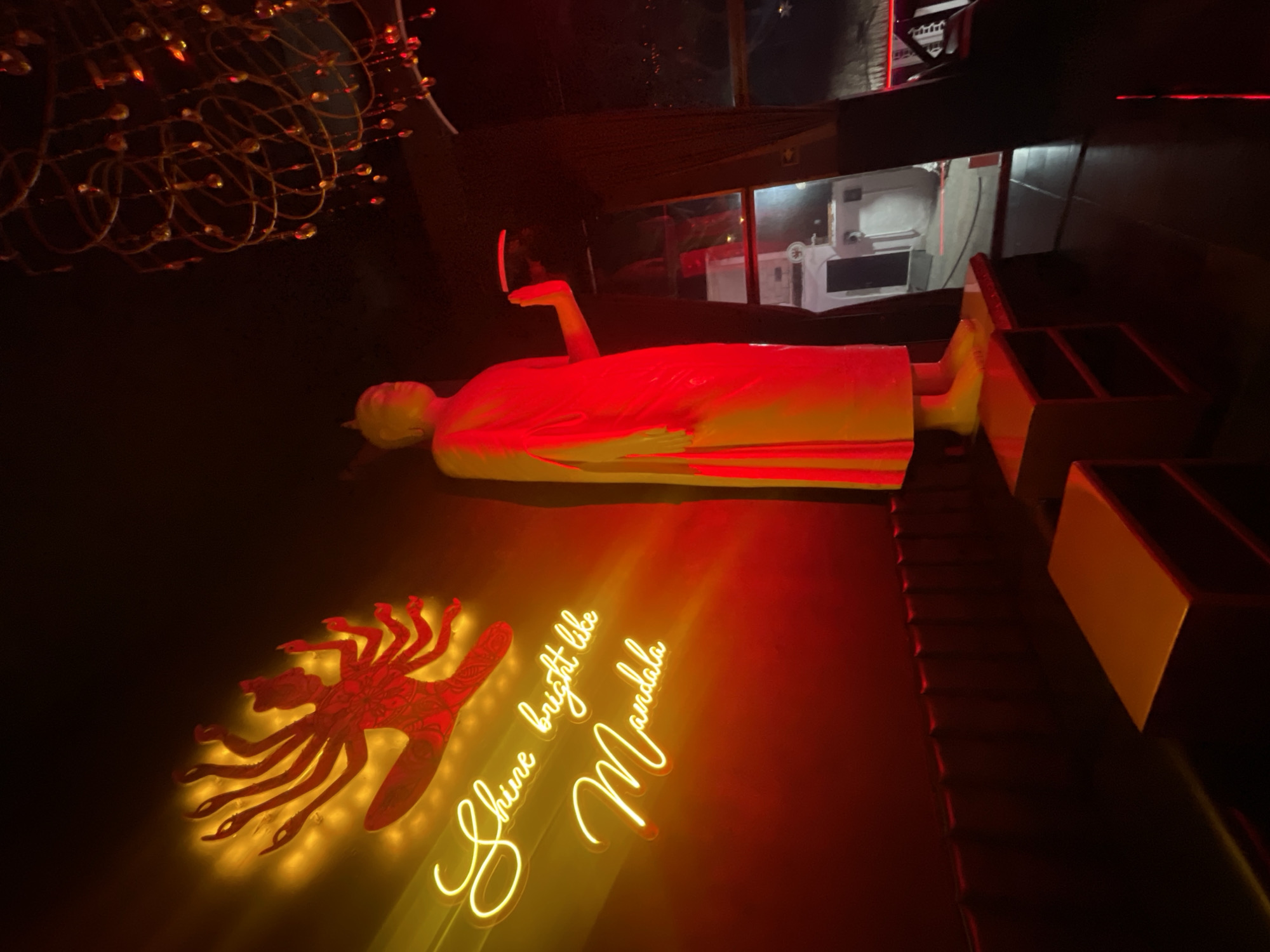 The image depicts an interior scene at an establishment in Playa del Carmen, Mexico. Dominating the foreground is a reclining Buddha statue, illuminated in red. A glowing neon sign reading "Shine bright like Mandala" accompanies a similarly lit red, hand-shaped design. To the right of the statue are several box-like structures, possibly seating or decorative elements. Overhead, a complex chandelier design adds to the ambient lighting. Through a window in the background, what appears to be a white interior with furniture or equipment is visible. The scene is dimly lit, contributing to a mysterious and atmospheric feel.