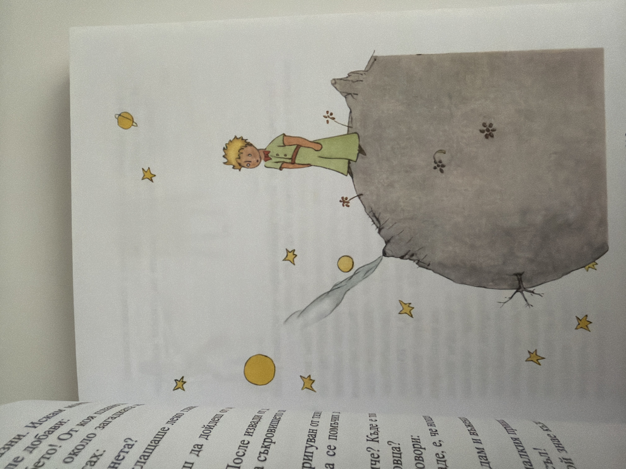 The image is a close-up of a page from a book, featuring an illustration from "The Little Prince." A young boy with blond hair, wearing a green suit and a red bow tie, stands on a barren, gray planet. He is looking off to the right. Several yellow stars and two small orange flowers are scattered around him and the planet. A faint gray plume, possibly representing smoke or a geyser, extends upwards from the planet's surface. The book page itself is white with black text visible on the left side, which appears to be in Bulgarian. The background of the image is a plain white surface, likely a table or floor. The image does not contain any information about the city of Pazardzhik, Bulgaria.