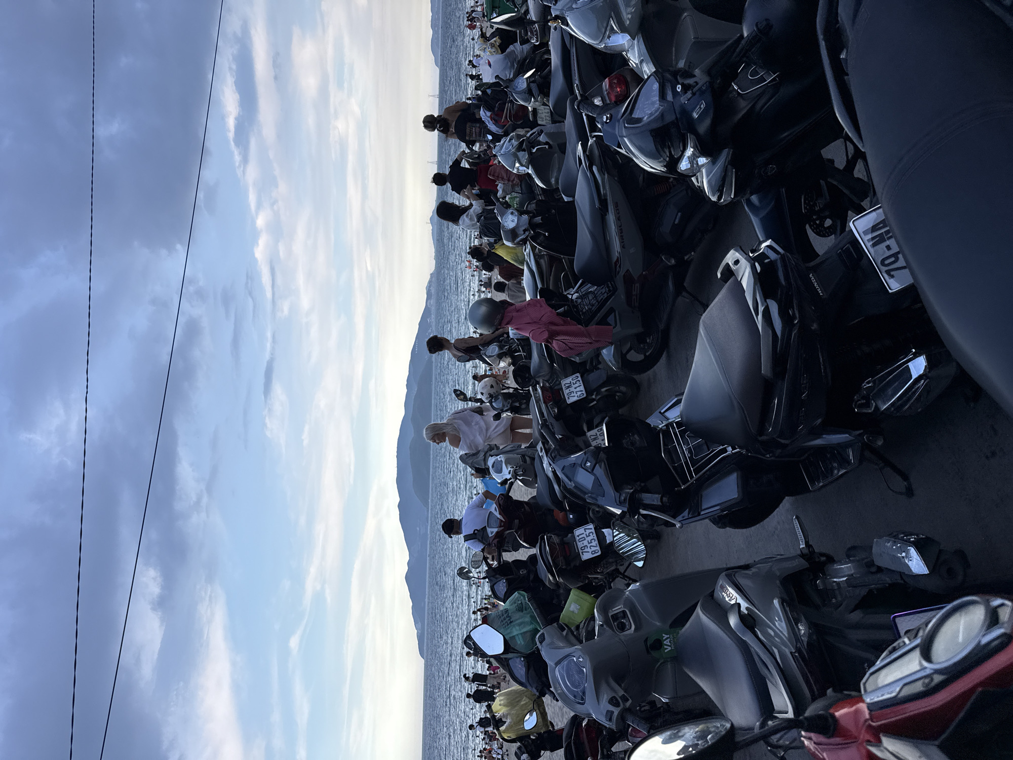 The image depicts a crowded beach scene in Nha Trang Ward, Vietnam, with a large number of parked motorbikes in the foreground. The beach itself is populated with many people, some of whom are standing near the water, while others are further back. Beyond the beach, the ocean stretches to the horizon, where islands are visible in the distance. The sky is partly cloudy, with a mix of blue and grey hues. The motorbikes, predominantly scooters, are tightly packed together, suggesting a popular location or event. The overall atmosphere appears to be lively and bustling, characteristic of a tourist area.