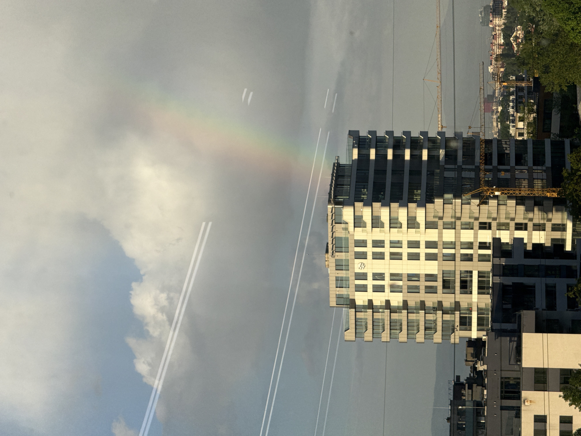 Here's a detailed description of the image:
**General Scene:**
The image is of a cityscape, seemingly taken from an elevated position (likely a high-rise building). It features a mix of modern architecture, construction elements, and natural phenomena. The orientation of the image is rotated 90 degrees counterclockwise.
**Specific Elements:**
* **Buildings:** A prominent, blocky, modern building with a grid-like facade is located in the middle of the frame. The building's windows create a pattern. Other buildings, including what appear to be residential structures, are visible in the background.
* **Construction:** There are construction cranes visible in the background, indicating ongoing development in the area.
* **Rainbow:** A faint but distinct rainbow is seen in the upper left of the image against a cloudy sky.
* **Atmosphere:** The sky is partly cloudy. The light suggests it is either early morning or late afternoon.
* **Lines:** Thin, near parallel lines vertically down the image, may be transmission lines.
**Location:**
While the exact street address cannot be determined solely from this image, the architecture, urban density, and presence of construction activity strongly suggest a location within Sofia, Bulgaria.
**People, Actions, Interactions:**
Given the wide-angle shot and lack of identifiable individuals, specific human actions or interactions cannot be discerned. The image primarily focuses on the built environment and the natural phenomenon of the rainbow.